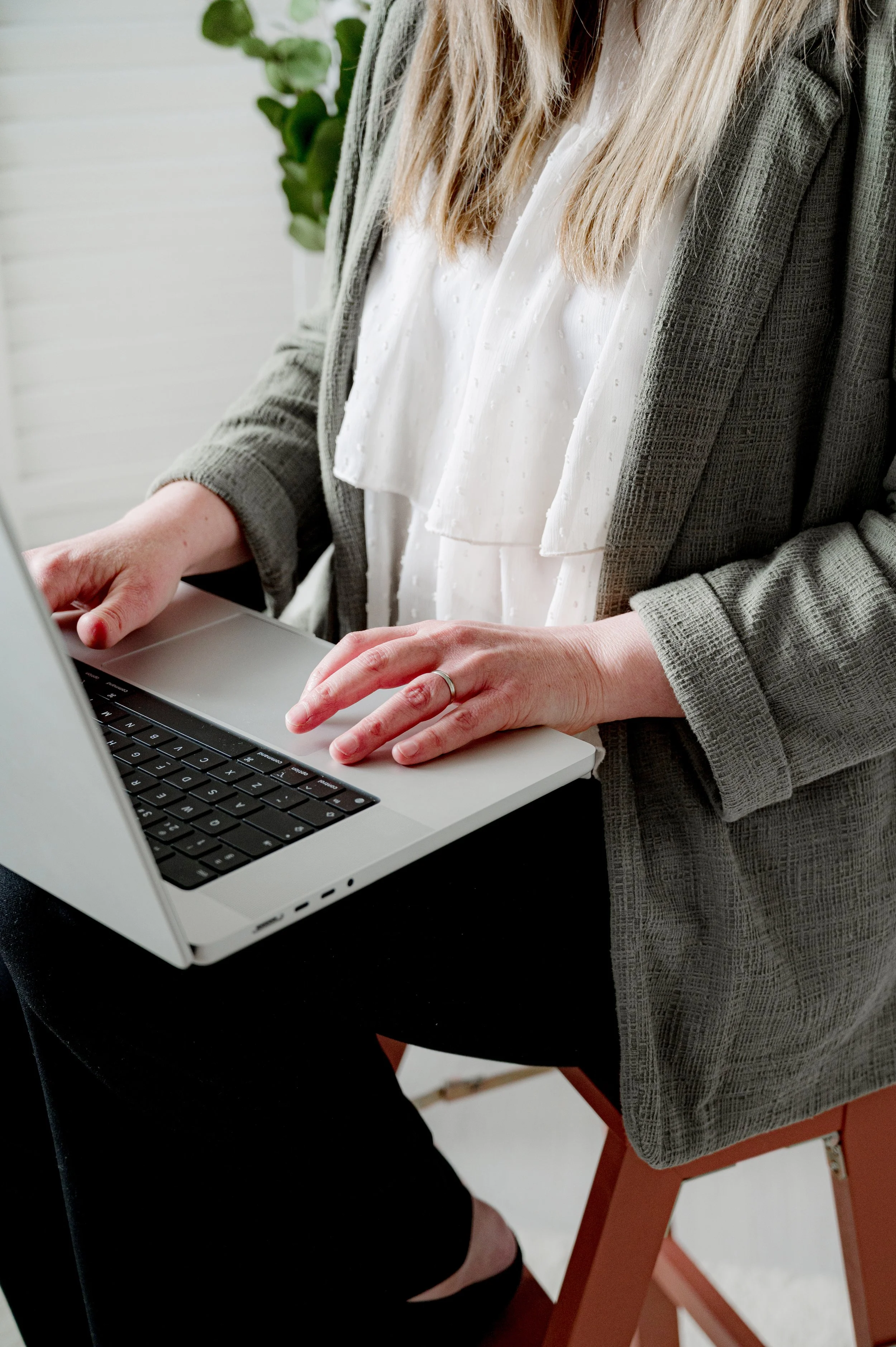 A woman with long blonde hair using a silver laptop, sitting on a red chair, wearing a white blouse with ruffles, a gray blazer, and black pants.