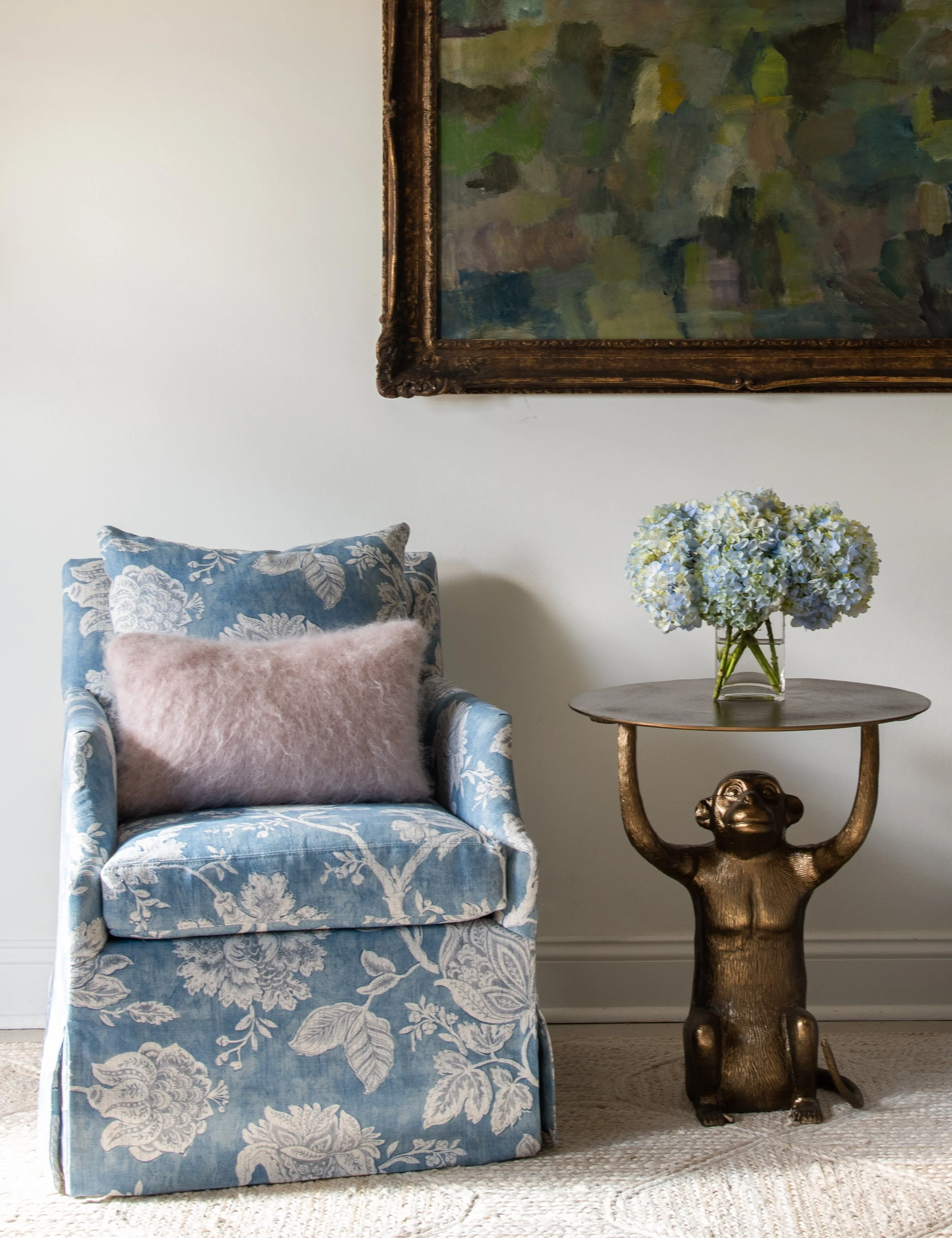 Blue, floral-patterned chair next to bronze side table beneath framed abstract painting.