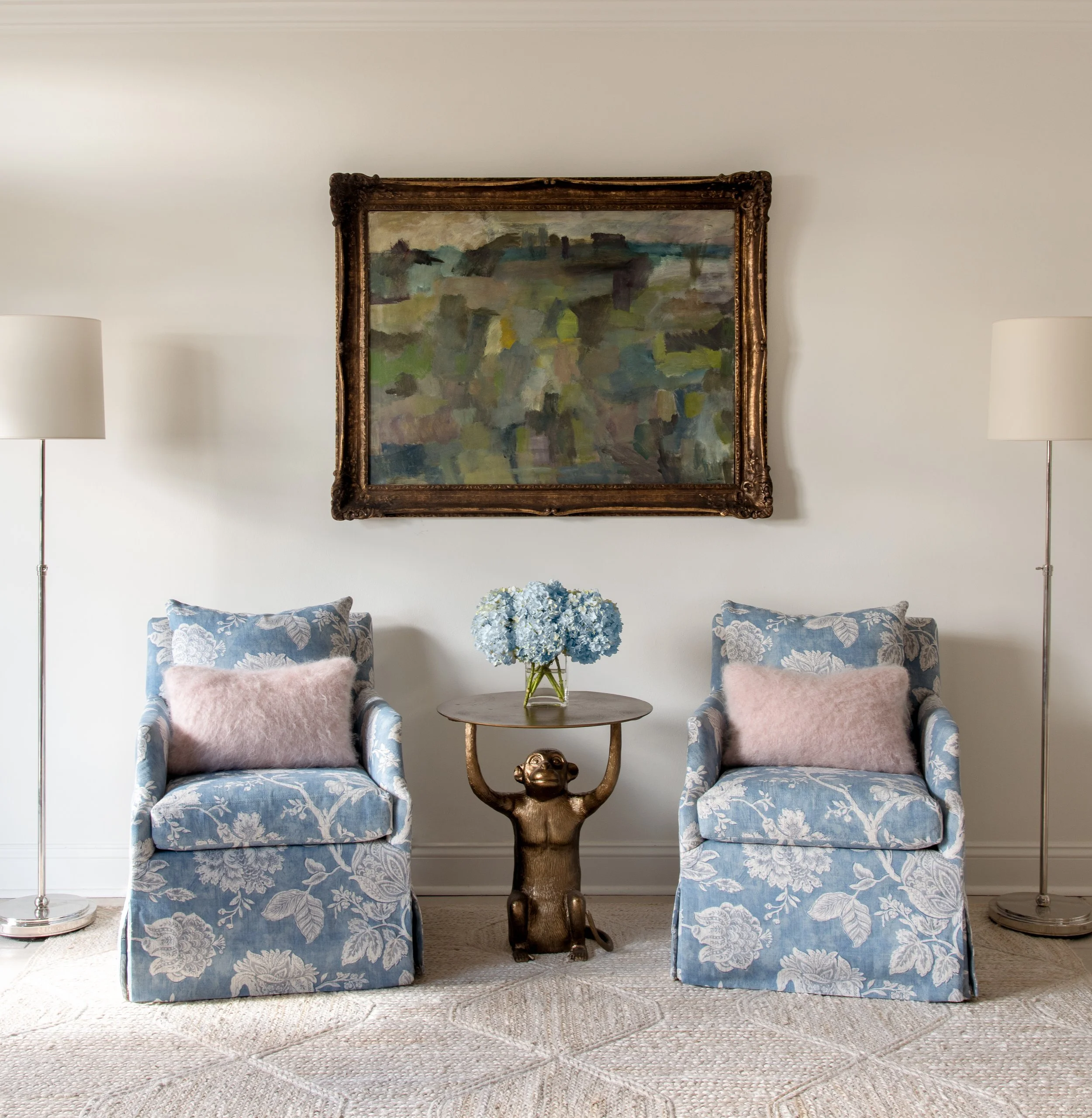Two blue, floral-patterned lounge chairs with bronze side table beneath abstract painting.