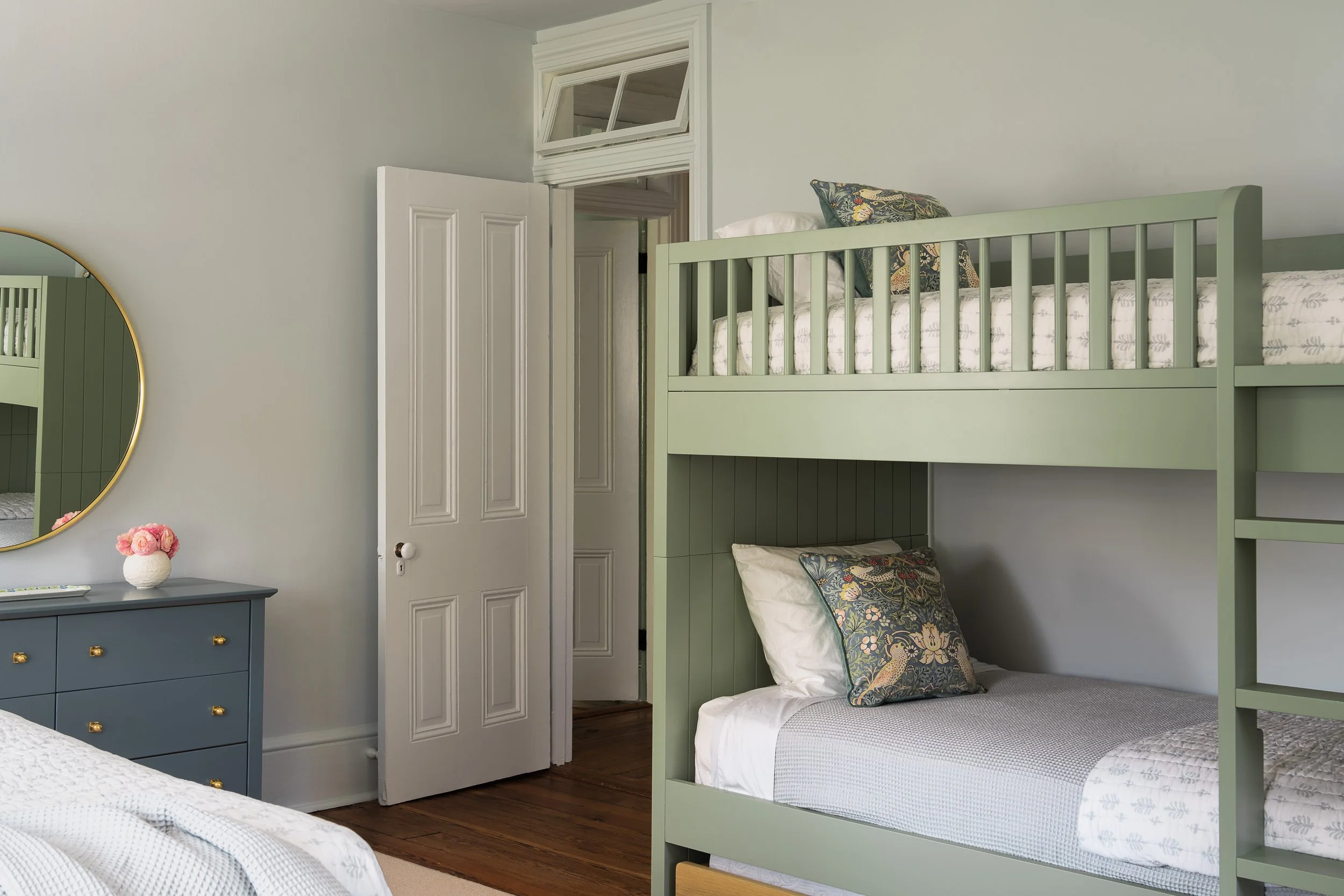 Bedroom with light green bunkbed, dark blue-green drawers and circular mirror.