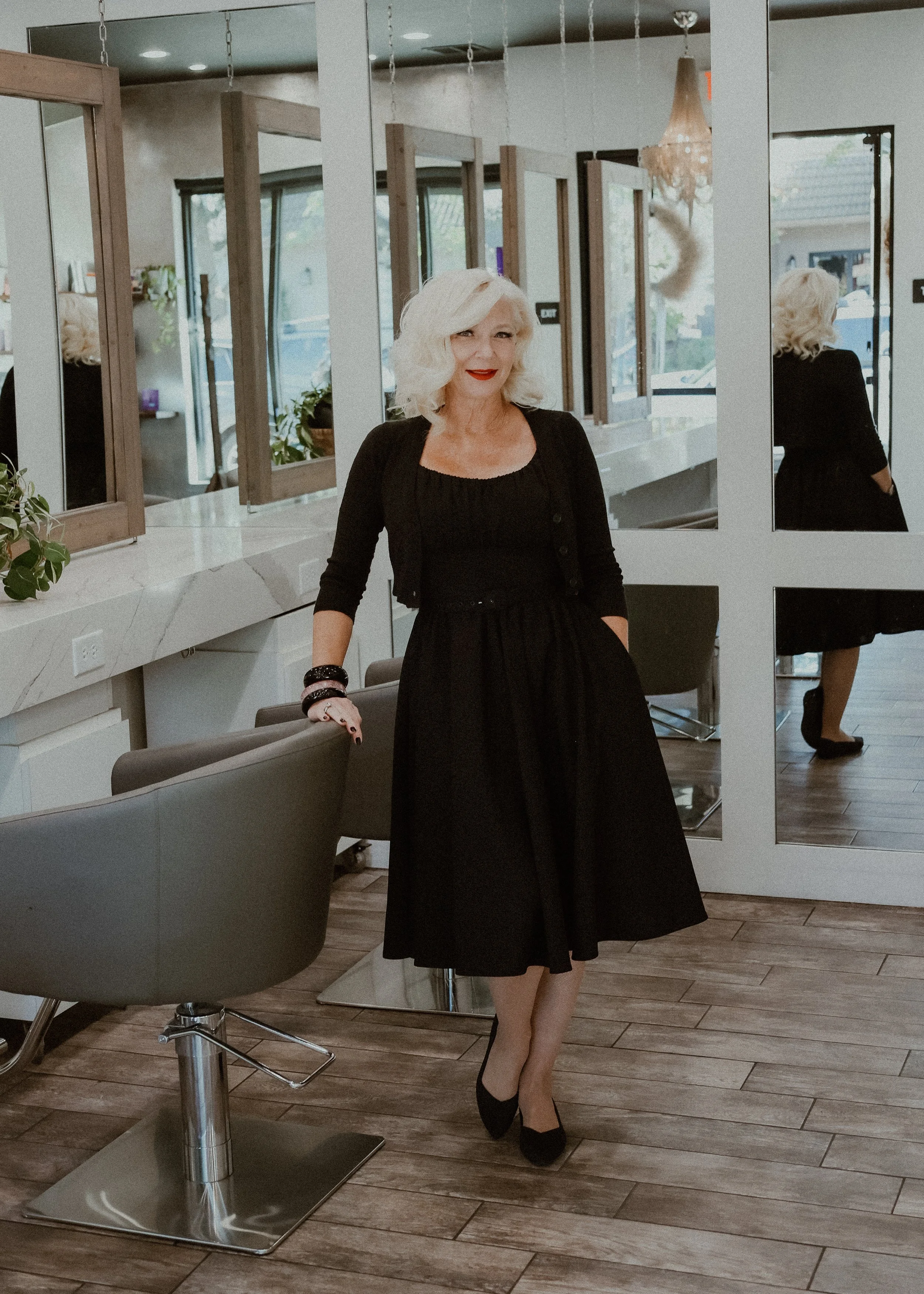 A woman with platinum blonde hair and red lipstick standing in a modern salon, wearing a black dress and heels, next to salon chairs and mirrors.