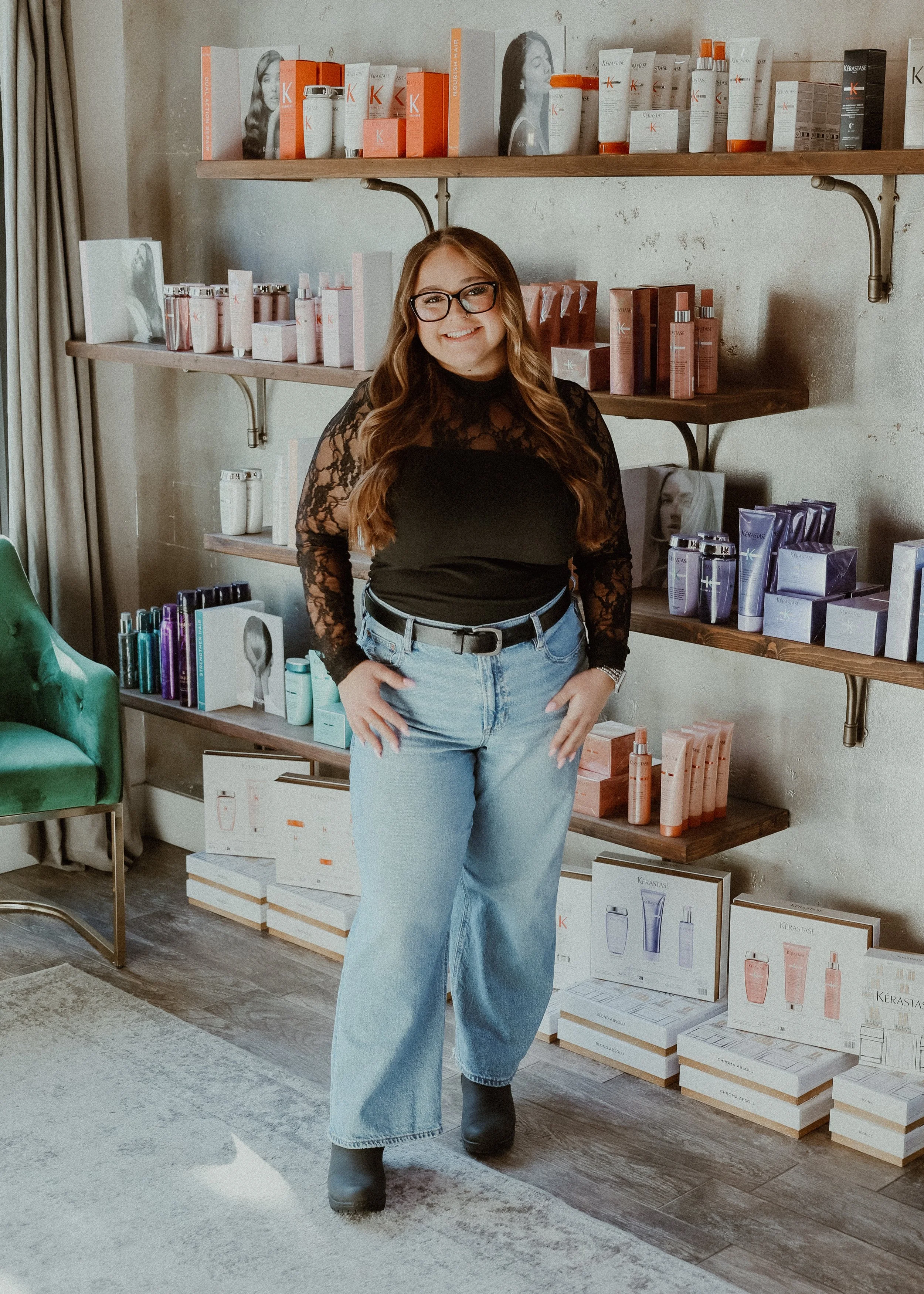 A woman with long brown hair, glasses, wearing a black lace top and blue jeans, standing in a beauty or skincare shop with shelves of cosmetic products behind her.