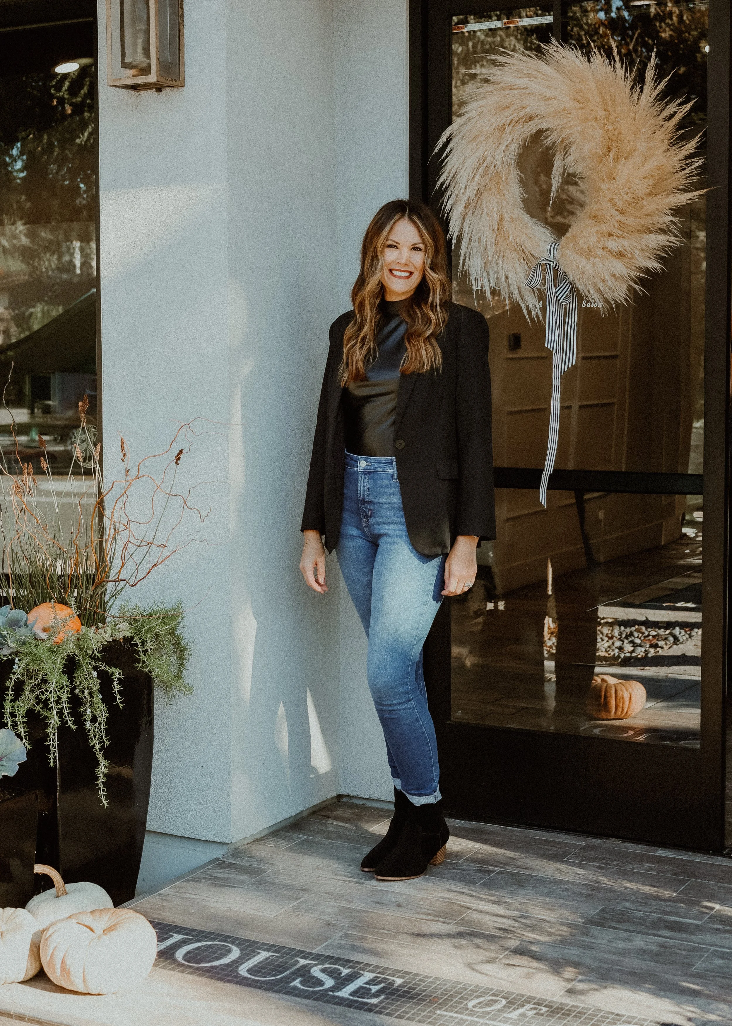 A woman with long wavy hair wearing a black blazer, black top, blue jeans, and black boots, standing outside a building decorated for fall with pumpkins and a large wreath on the door.