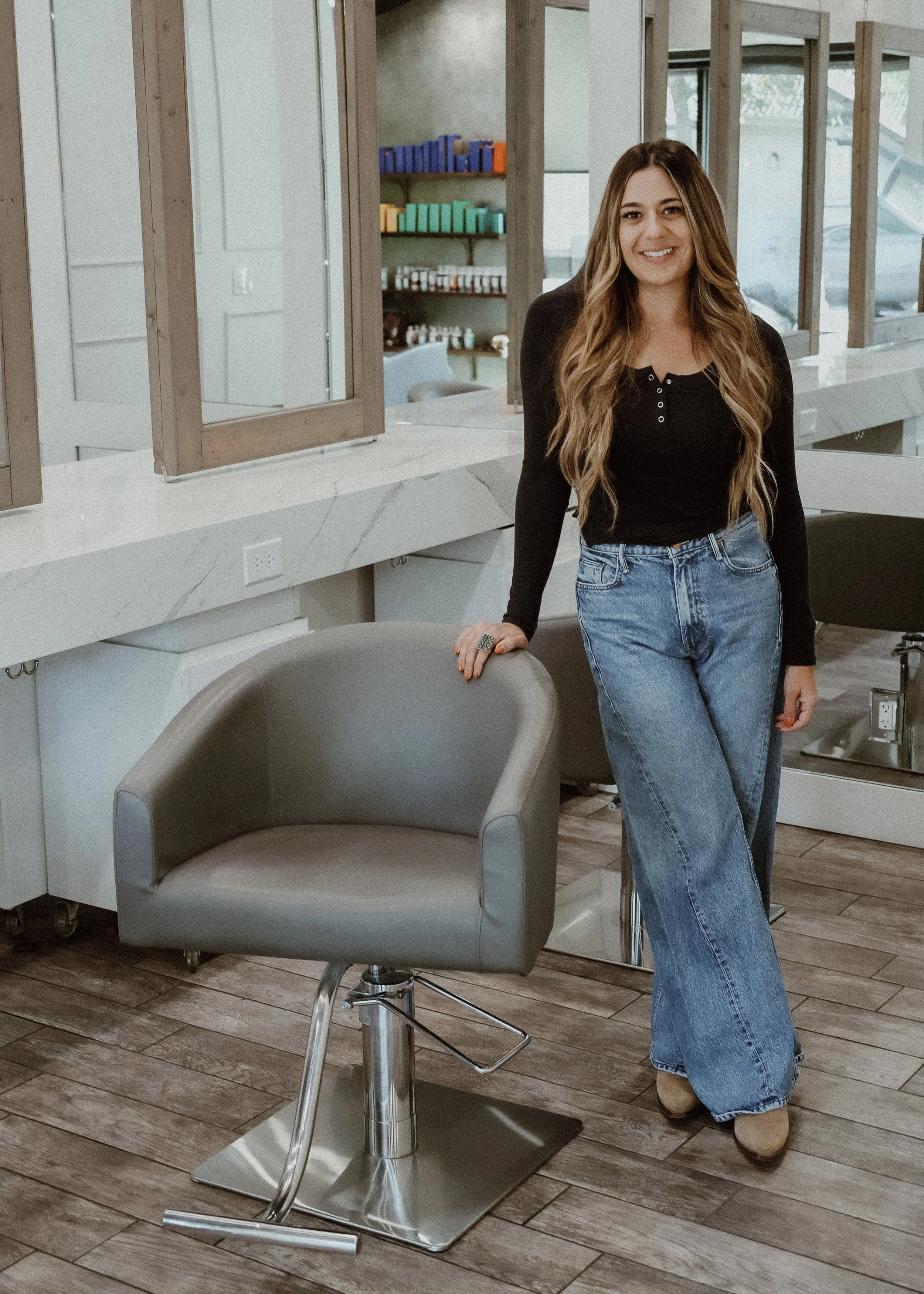A young woman with long wavy hair, wearing a black long-sleeve shirt and blue jeans, stands in a modern salon or studio, leaning on a gray salon chair with a wooden floor and shelves in the background.