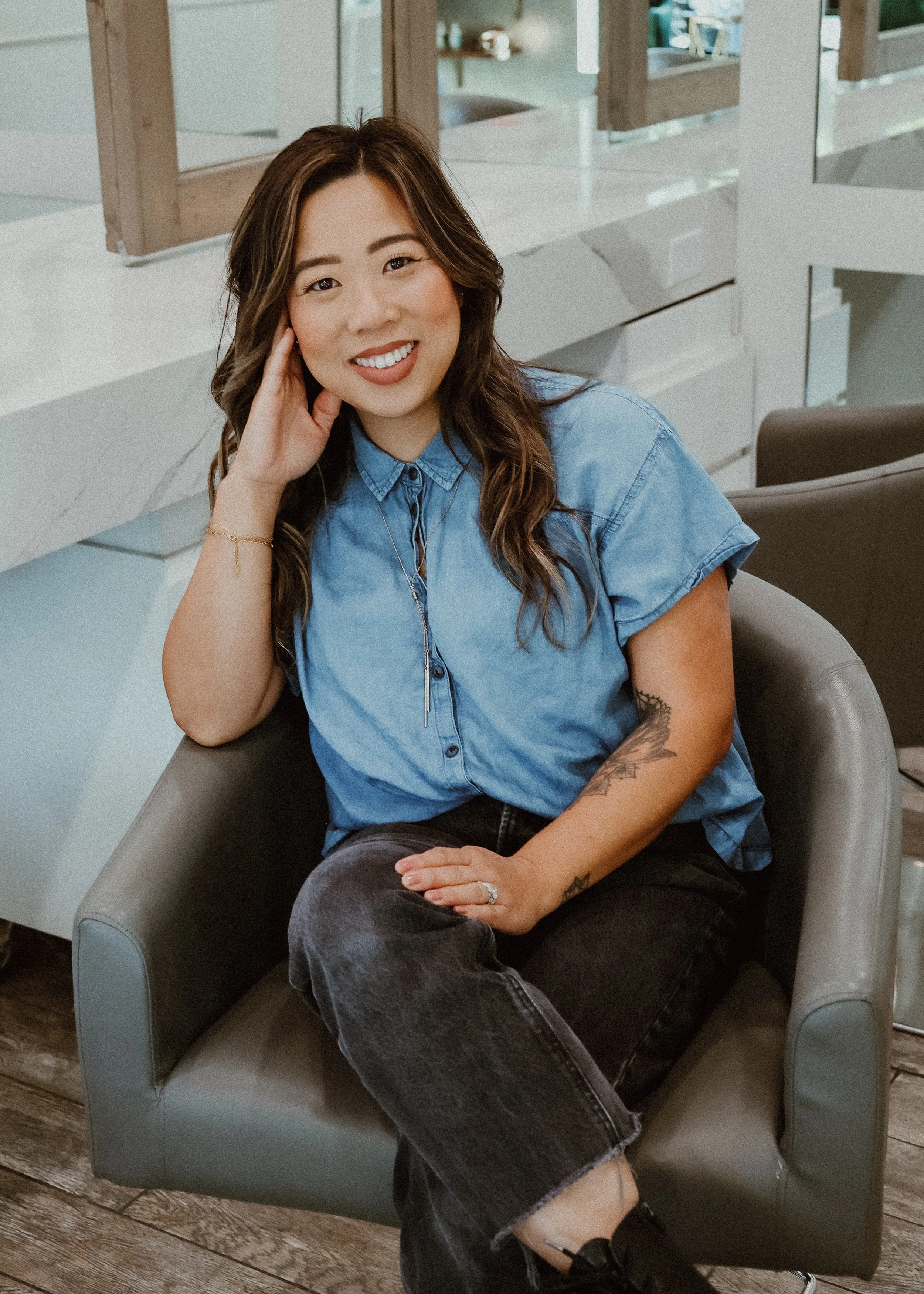 A woman sitting in a chair, smiling, with her left hand resting on her head. She is wearing a blue denim shirt, black jeans, and has tattoos on her right arm and left hand.