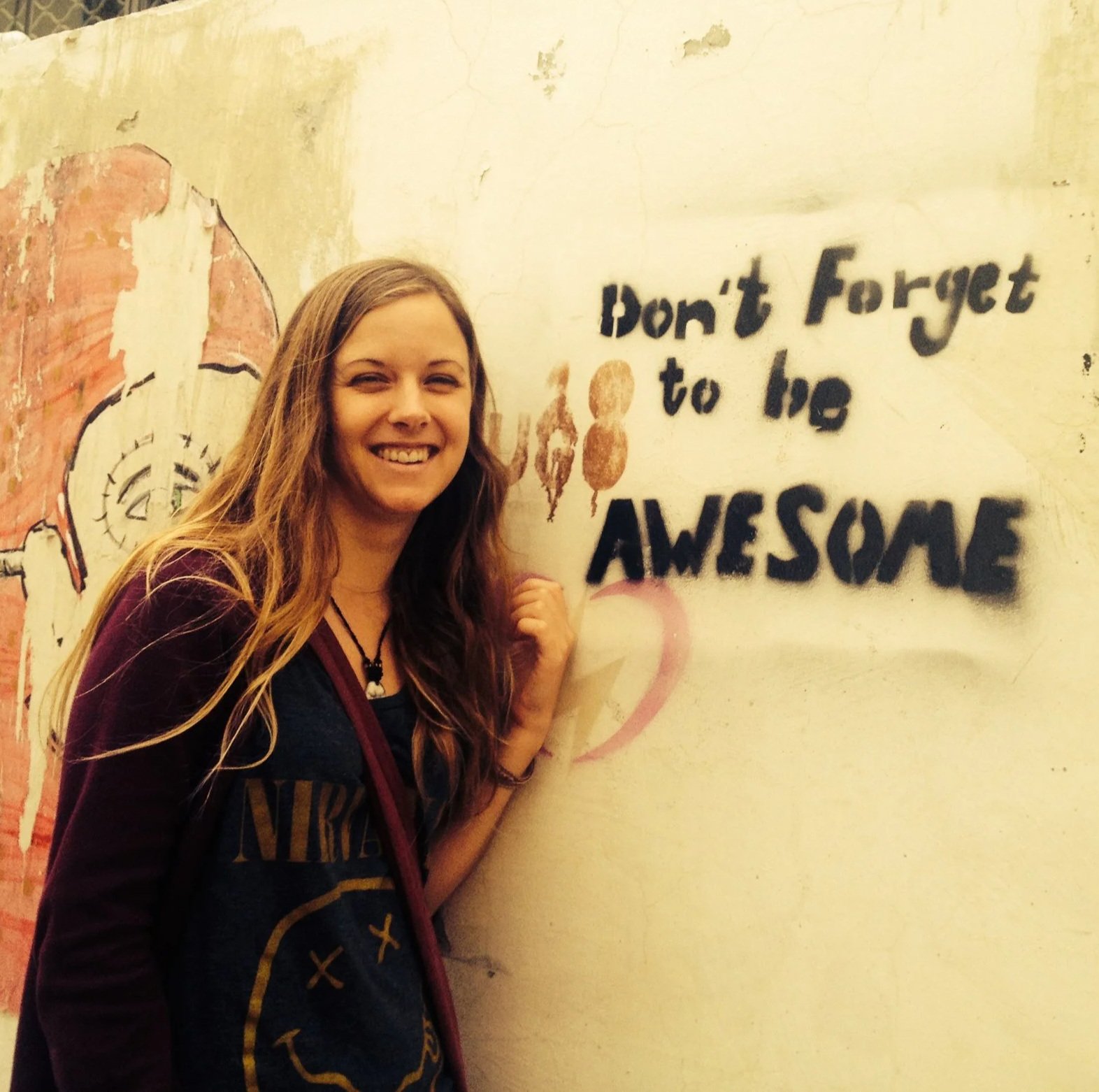A smiling young woman with long wavy hair standing in front of a wall with graffiti that says "Don't Forget to be AMAZING," next to a red heart shape.