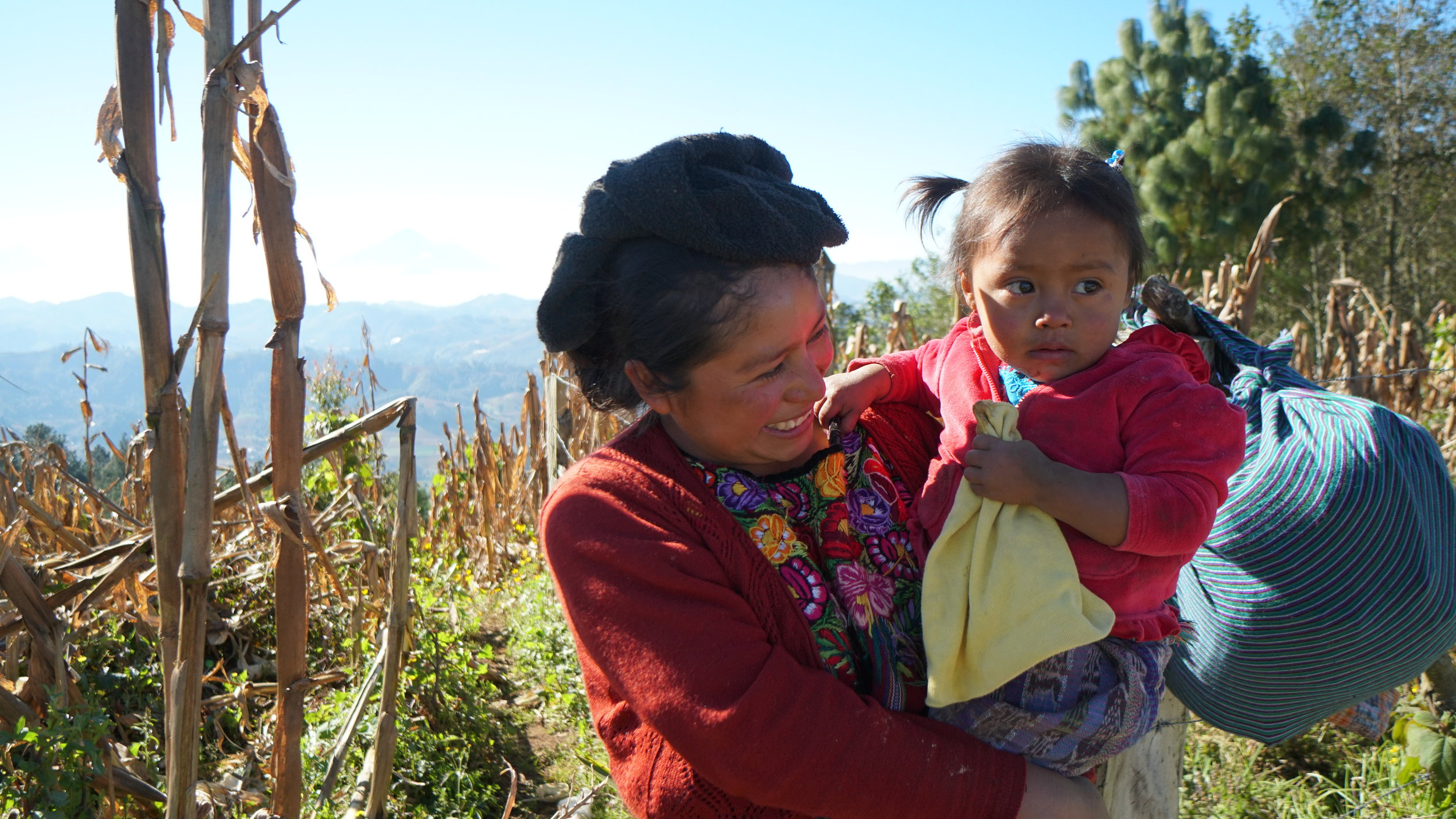 An indigenous woman dressed in a red sweater and flowered dress holding her young daughter