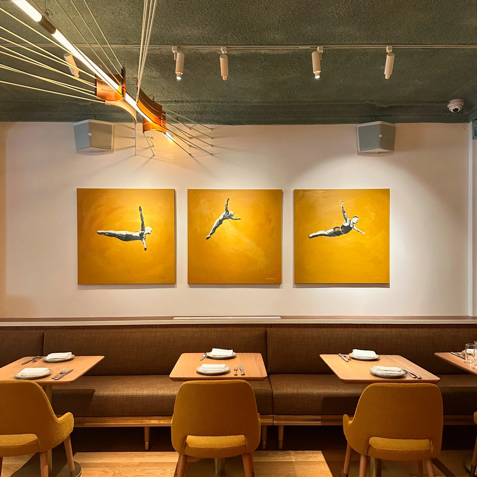 A restaurant dining area with a brown upholstered bench, four tables set with white plates and silverware, yellow chairs, and three large paintings of divers against a yellow background on the wall.