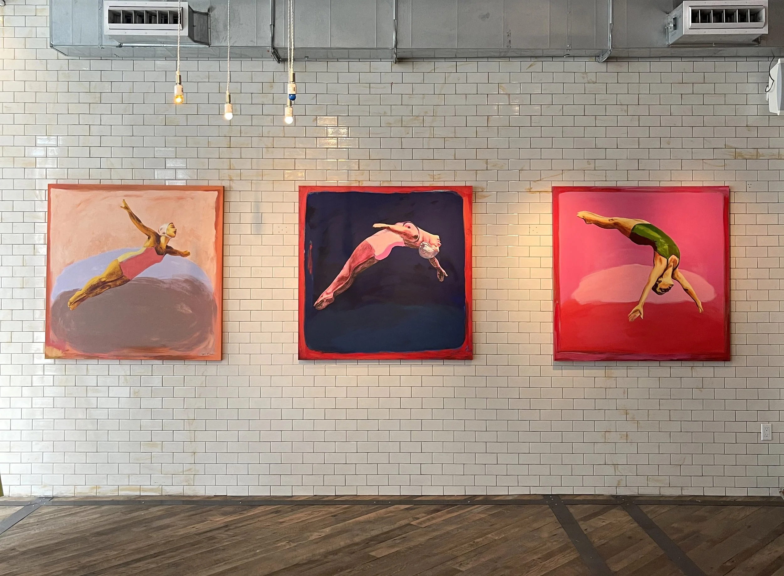 Three colorful paintings of swimmers diving into water, hanging on a white brick wall in an art gallery.