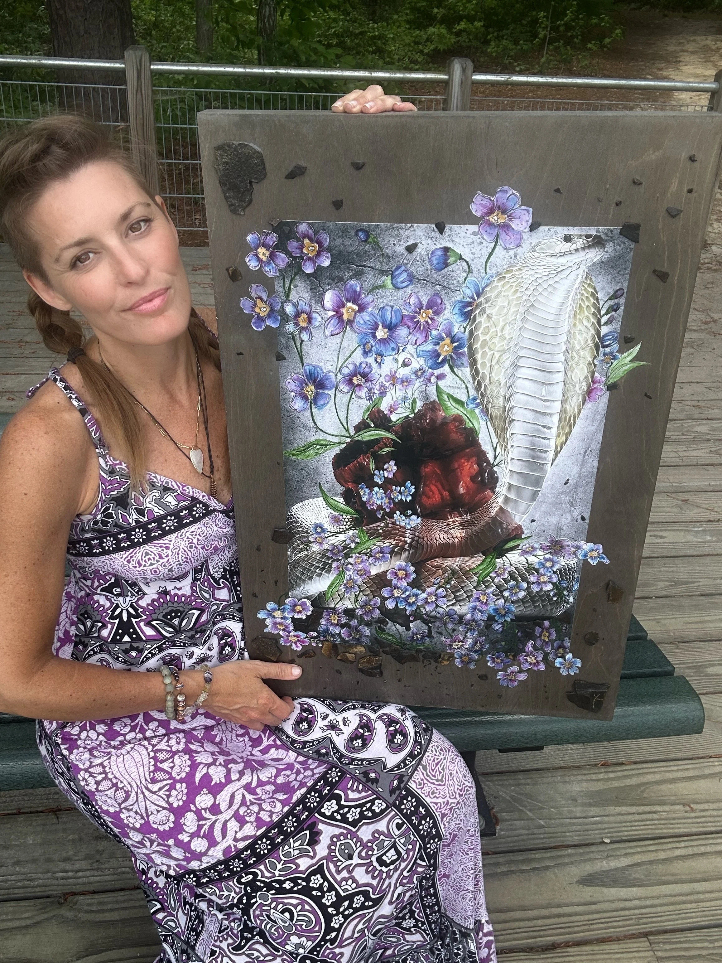 Artist Yuli Anun holding a piece of artwork. Her hair is braided. She is seated with a purple patterned dress. The art is mixed media with a cobra snake, anatomical heart, flowers, and rocks.