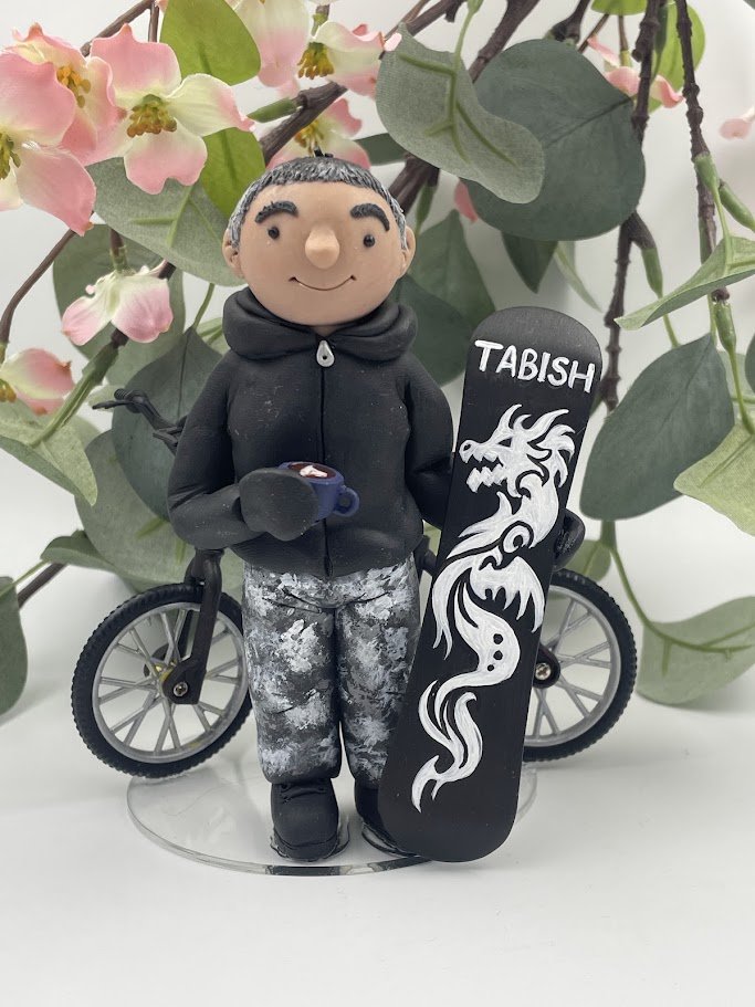 A polymer clay birthday cake topper of a man in a black hoddie and grey trousers holding a snowboard that says 'Tabish' with a dragon on it, holding a cup of coffee with a mountain bike behind