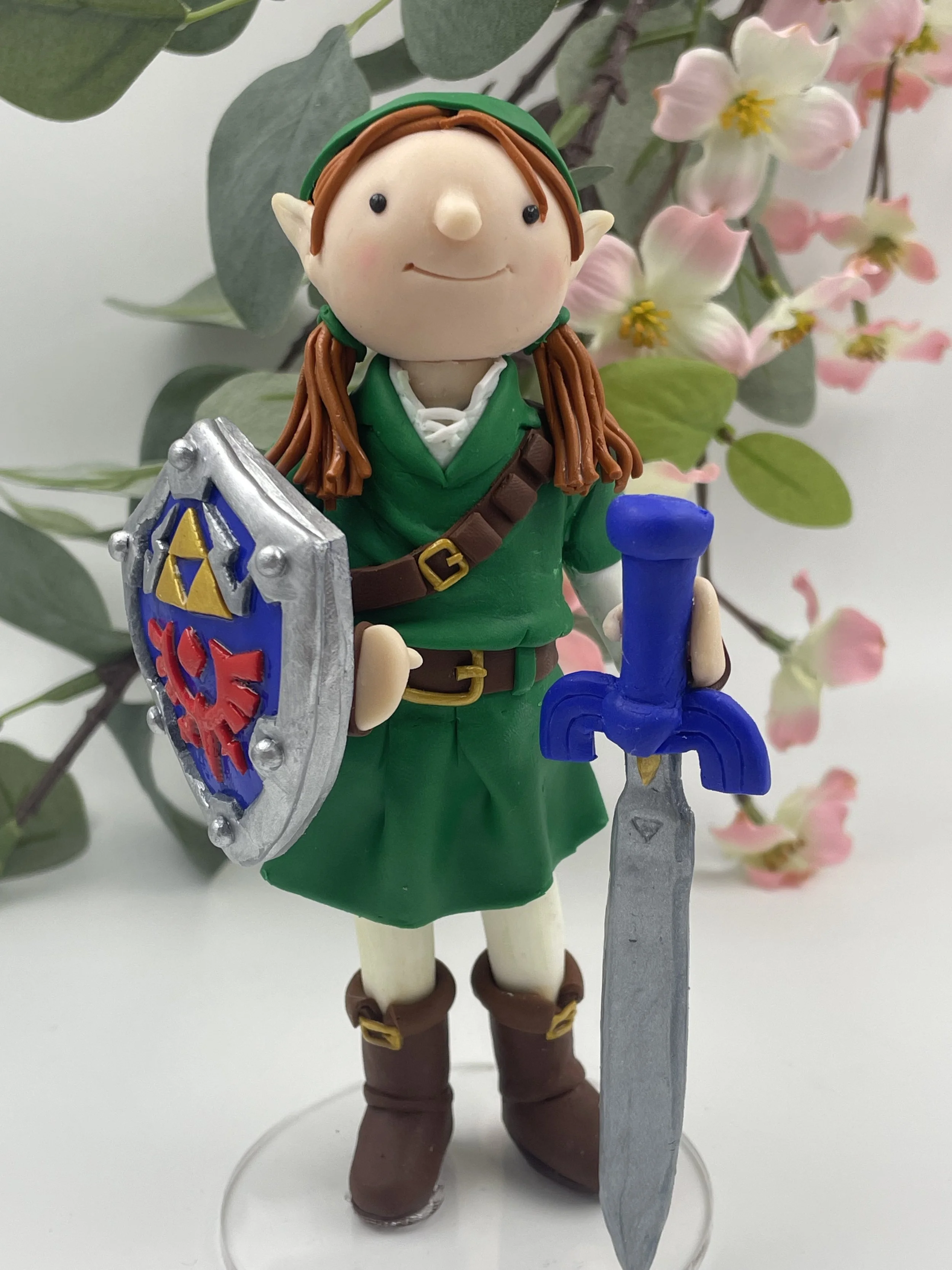 A polymer clay birthday cake topper of a red haired girl with bunches dressed as Link from The Legend of Zelda video game, holding a sword and shield