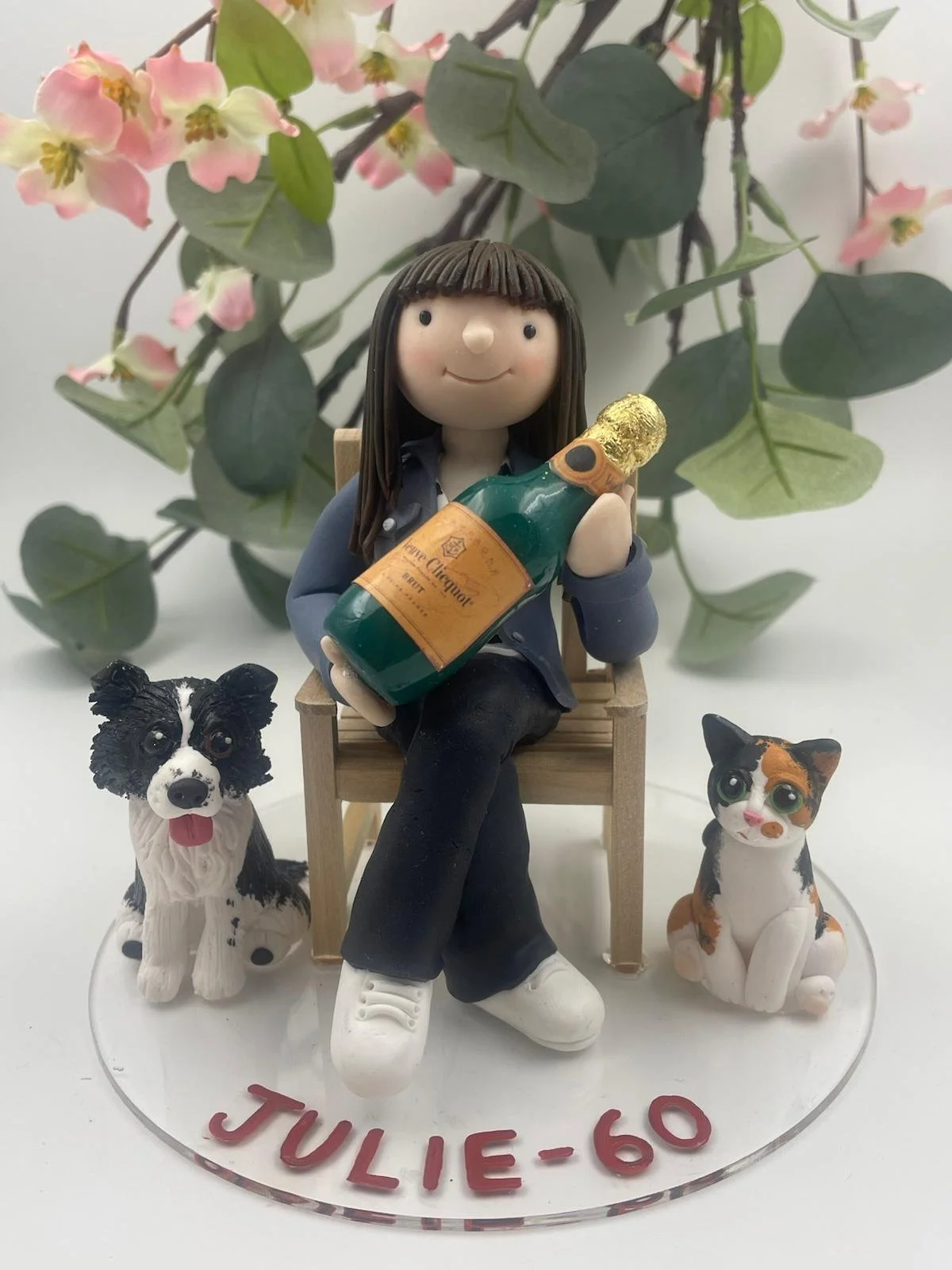 A clay 60th birthday cake topper of a woman with brown hair sitting in a garden chair with a magnum of champagne, with a dog and cat sitting beside her. It says 'Julie-60' in red around the perspex base.