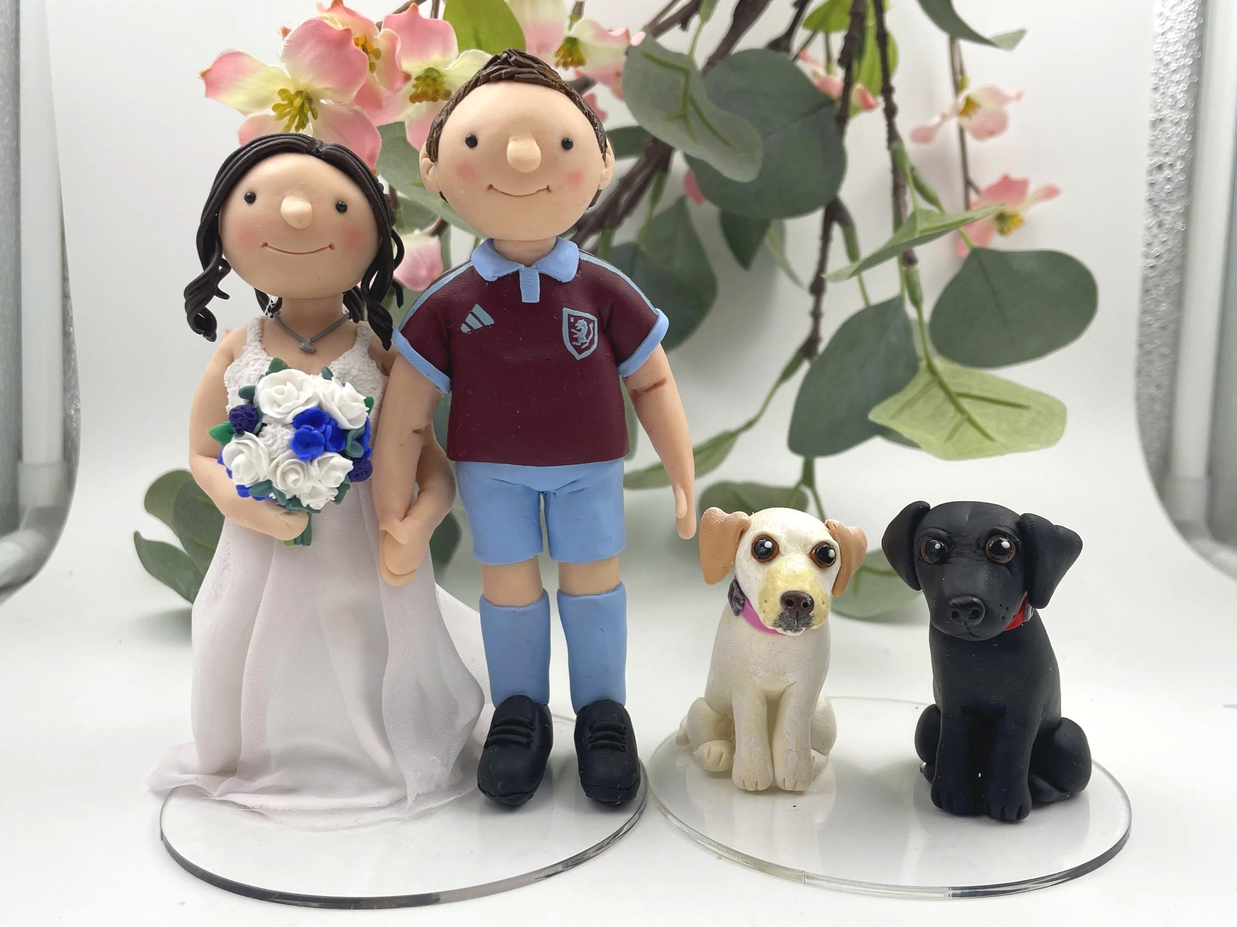 A polymer clay wedding cake topper of a bride in a whilte dress with a white and blue bouquet and the groom in a West Ham football strip, and two labrador retrievers, one white and one golden, on a white background with pink flowers