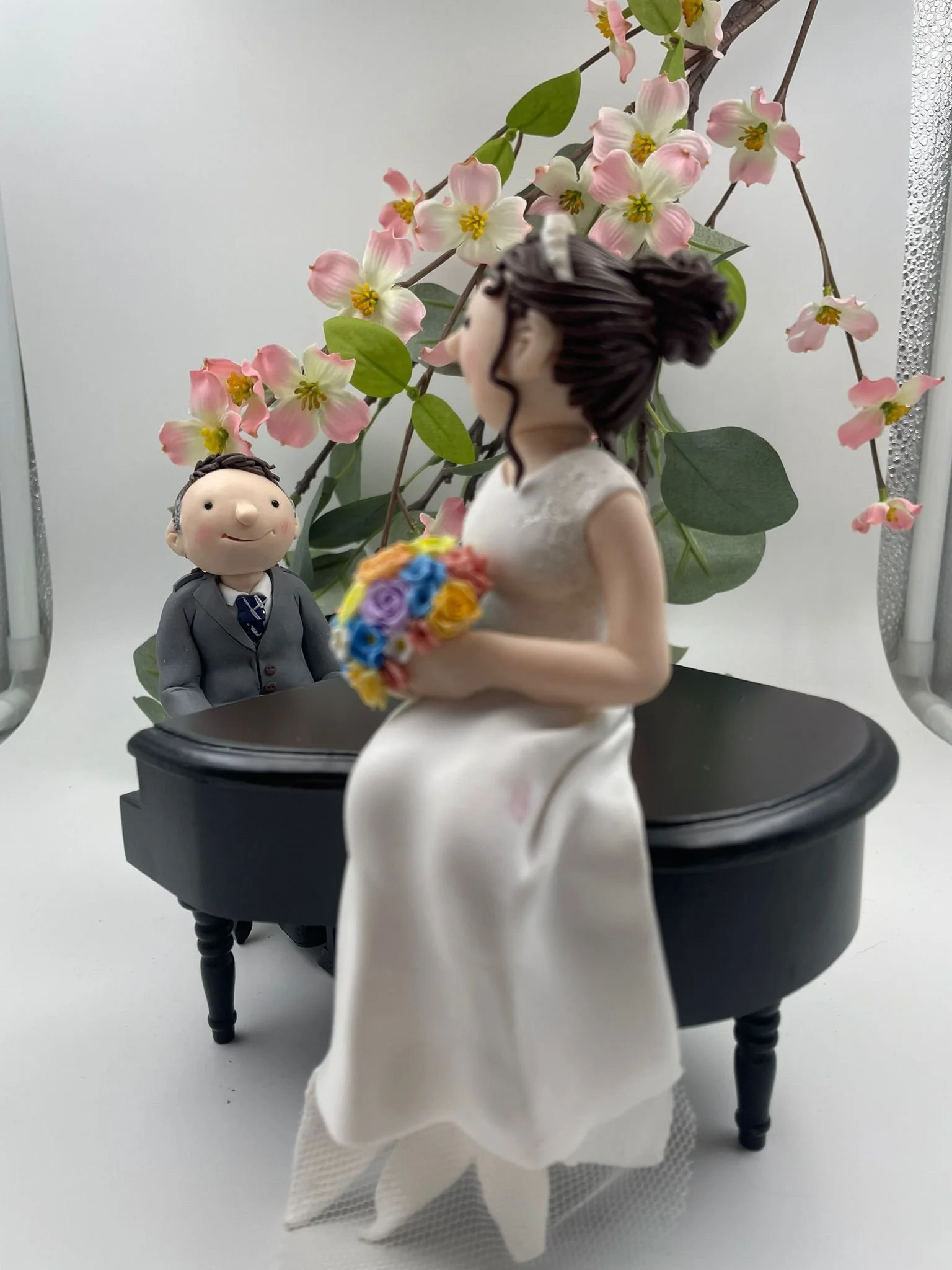 A polymer clay wedding cake topper featuring a bride sitting on a grand piano and the groom playing it