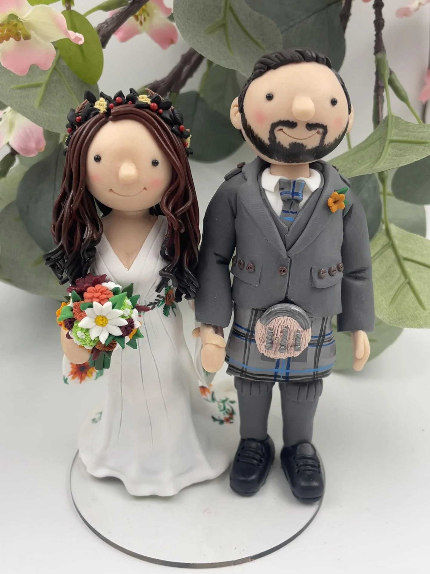 A polymer clay wedding cake topper of a bride in an autumnal headdress with an embroidered white dress and a bouquet of orange and red flowers, and a groom in a kilt