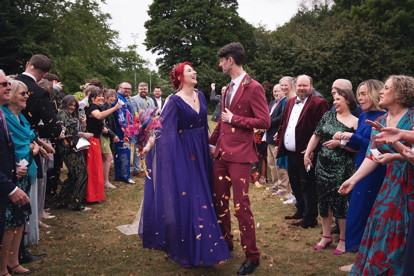A bride in a purple dress and a groom in a red suit surrounded by guests throwing confetti