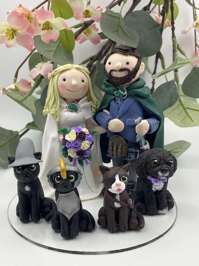 A polymer clay wedding cake topper featuring a bride and groom in Lord of the Rings-themed fantasy outfits, and their four pets wearing hats and outfits
