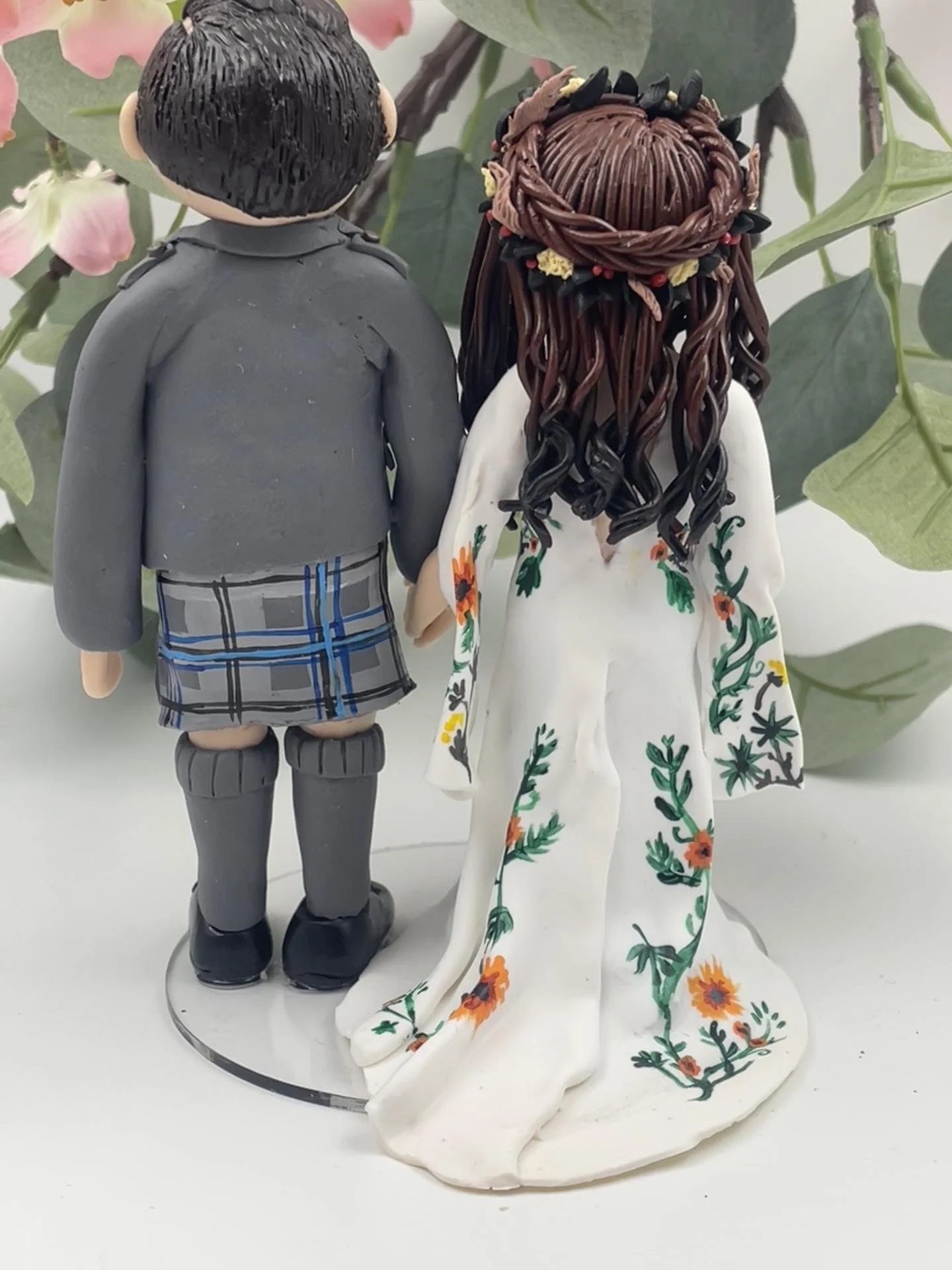 A polymer clay wedding cake topper of a bride in an autumnal headdress with an embroidered white dress and a bouquet of orange and red flowers, and a groom in a kilt