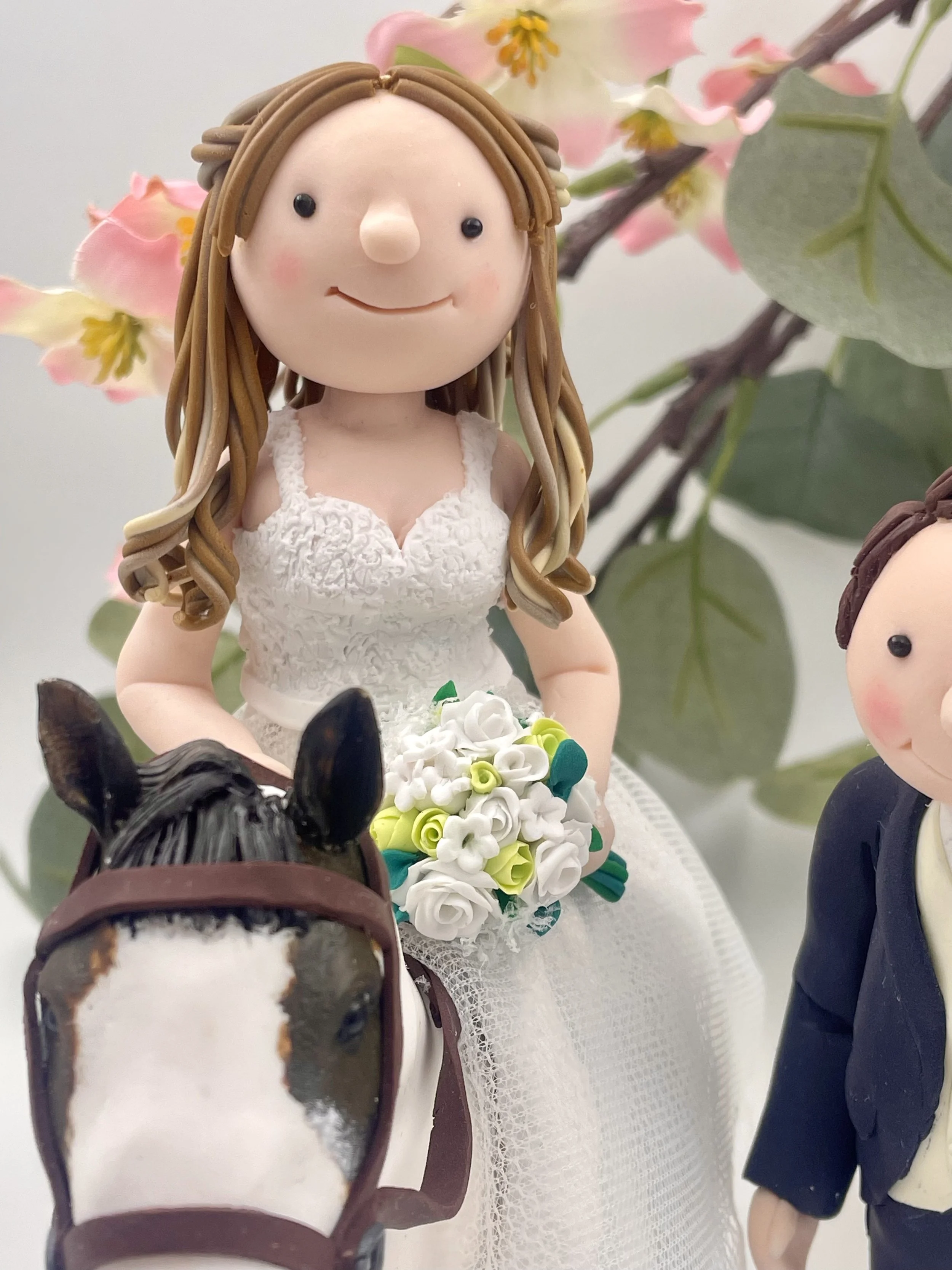 A polymer clay wedding cake topper of a blonde-haired bride riding a white and brown horse, against a background of pink flowers