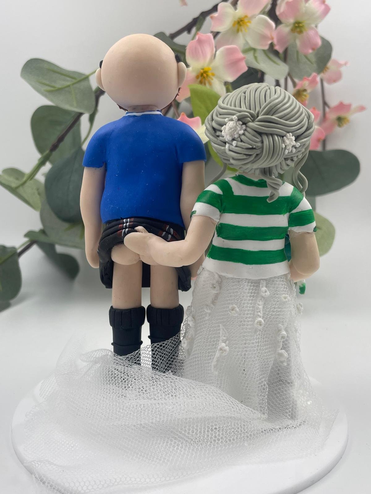 A wedding cake topper of a bride and groom in Celtic and Glasgow Rangers foodball shirts, the birde is lifting the groom's kilt at the back