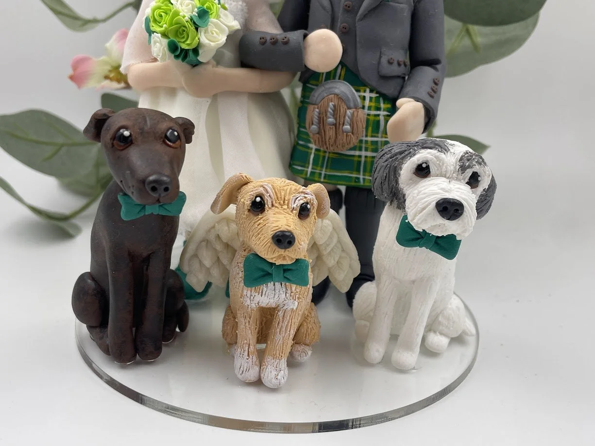 A close up of a handmade clay cake topper featuring three dogs in green bowties, one of whom has angel wings