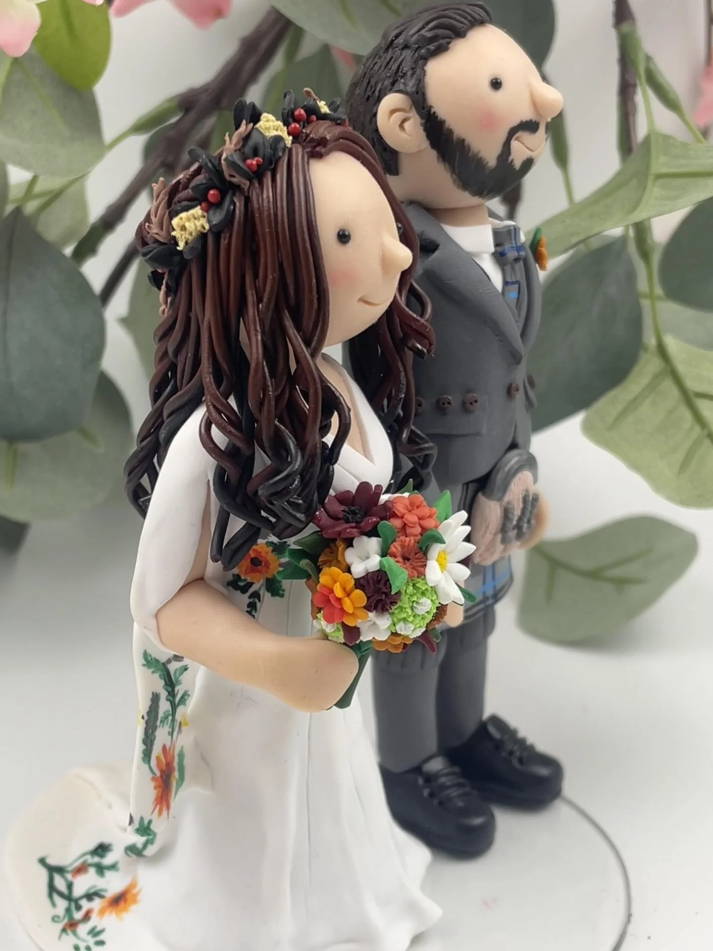 A polymer clay wedding cake topper of a red-haired bride in an autumnal headdress with an embroidered white dress and a bouquet of orange and red flowers, and a groom in a kilt