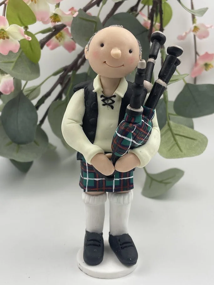 A polymer clay birthday cake topper of a man in a kilt and ghillie shirt with a set of bagpipes