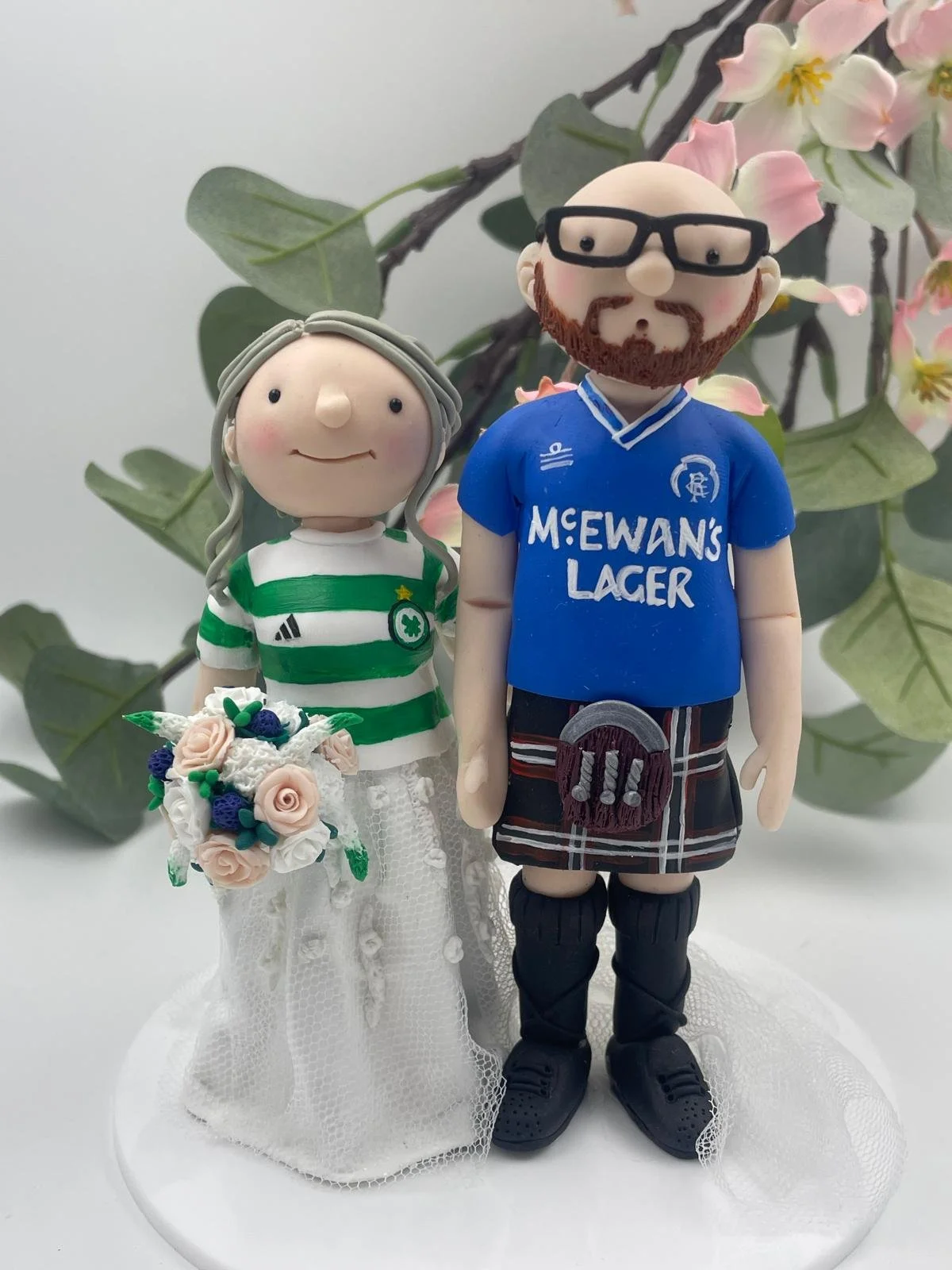 A wedding cake fopper featuring a bride and groom wearing Glasgow Rangers and Celtic football shirts. The bride is lifting the groom's kilt