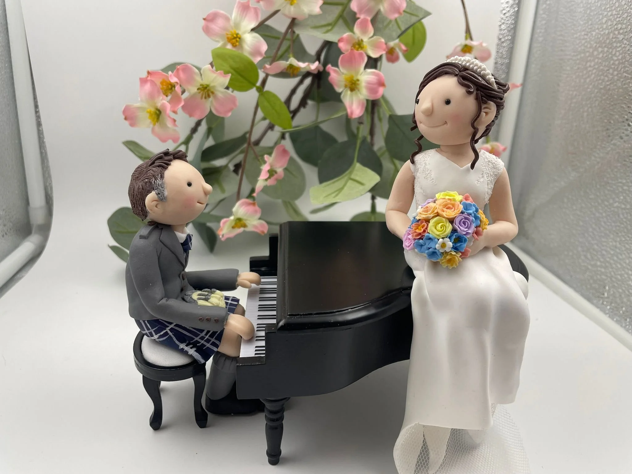 A polymer clay wedding cake topper featuring a bride sitting on a grand piano and the groom playing it