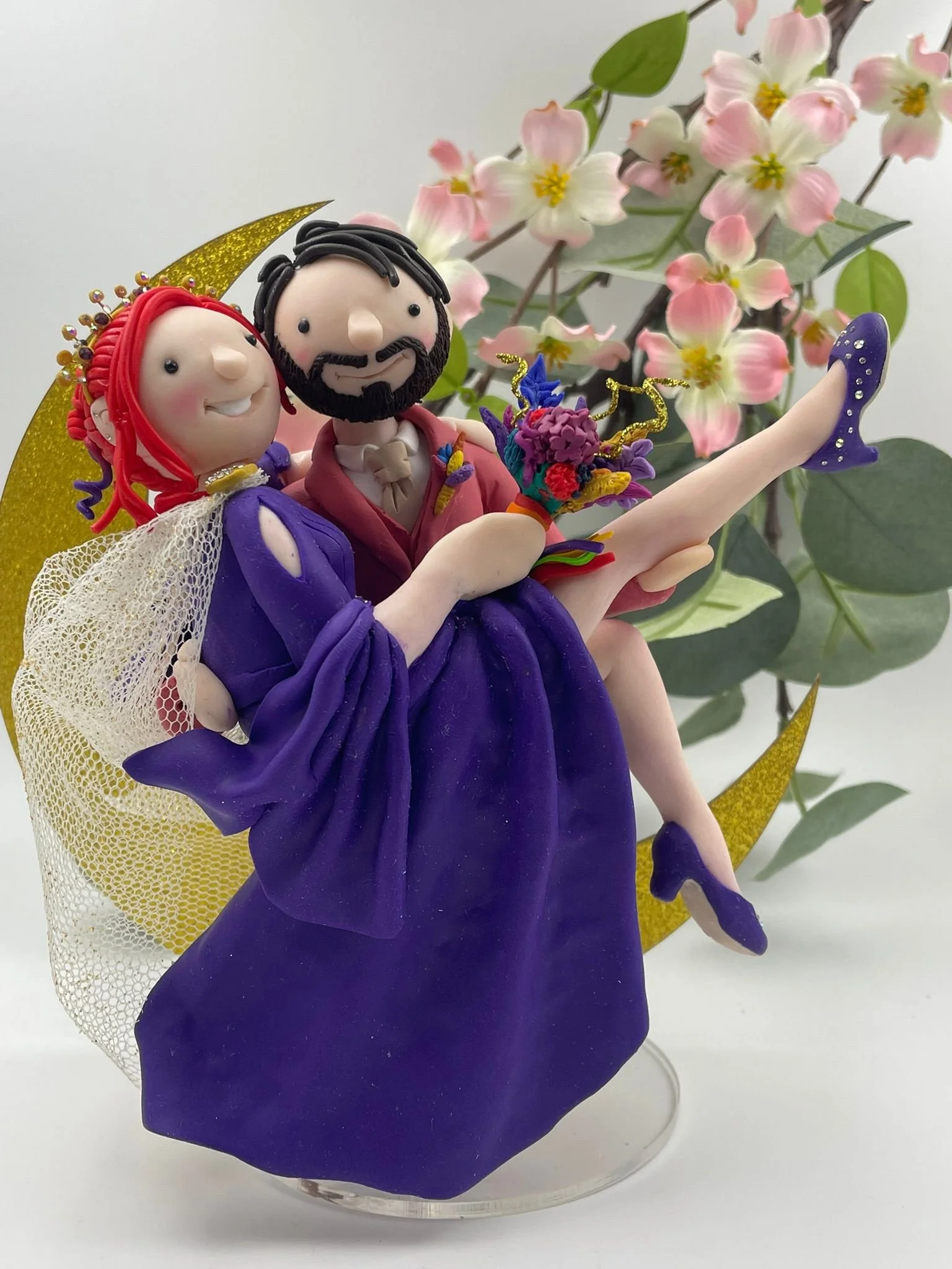 a polymer clay wedding cake topper featuring a bride in a purple dress and a groom in a pink suit in a romantic pose, with a gold crescent moon behind