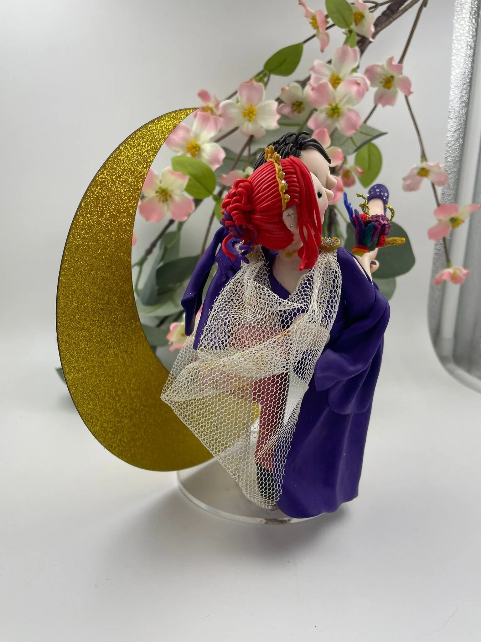a polymer clay wedding cake topper featuring a bride in a purple dress and a groom in a pink suit in a romantic pose, with a gold crescent moon behind