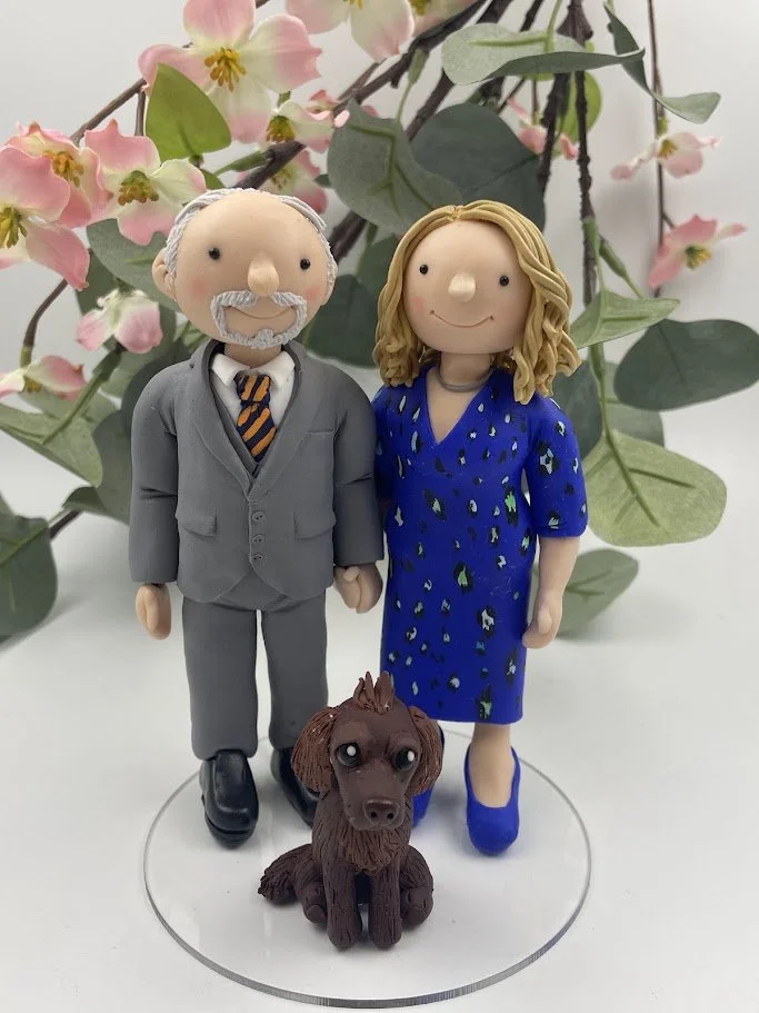 A polymer clay wedding anniversary cake topper of a couple in a grey suit and a blue dress with a brown spaniel dog
