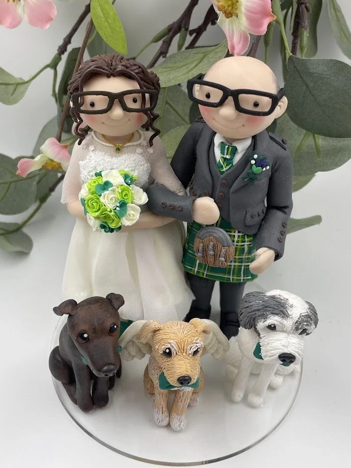A clay wedding cake topper of a bride and groom in glasses with three dogs surrounding them