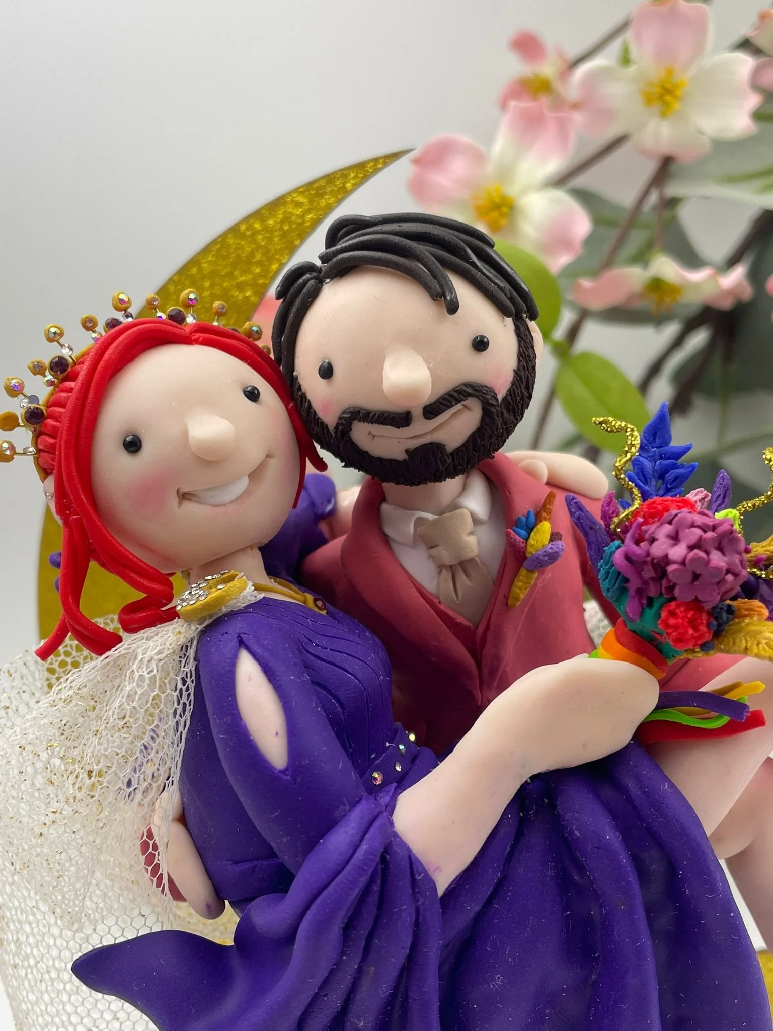 Close up of a polymer clay wedding cake topper featuring a bride in a purple dress and a groom in a pink suit in a romantic pose, with a gold crescent moon behind