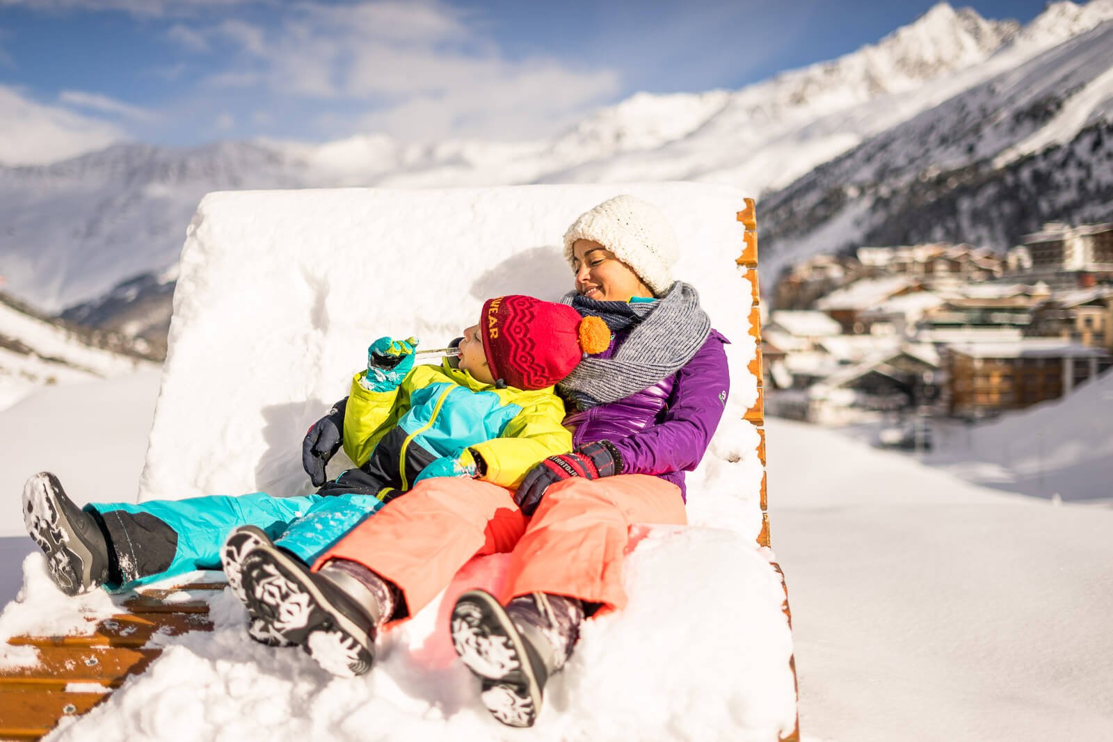 Mutter und Sohn entspannen auf einer Schneeliege vor einem Winterhotel, sonniger Tag in den Alpen.