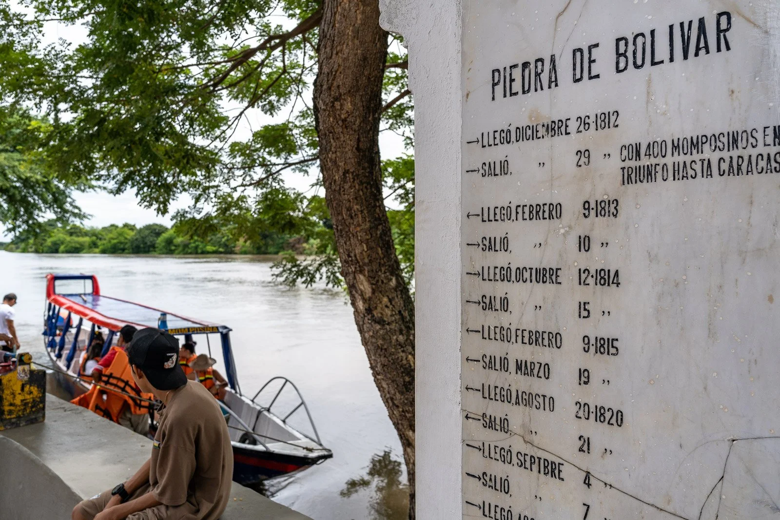  Gedenktafel „Piedra de Bolívar“ am Río Magdalena in Mompox, mit vorbeifahrendem Boot und wartenden Passagieren am Ufer. 