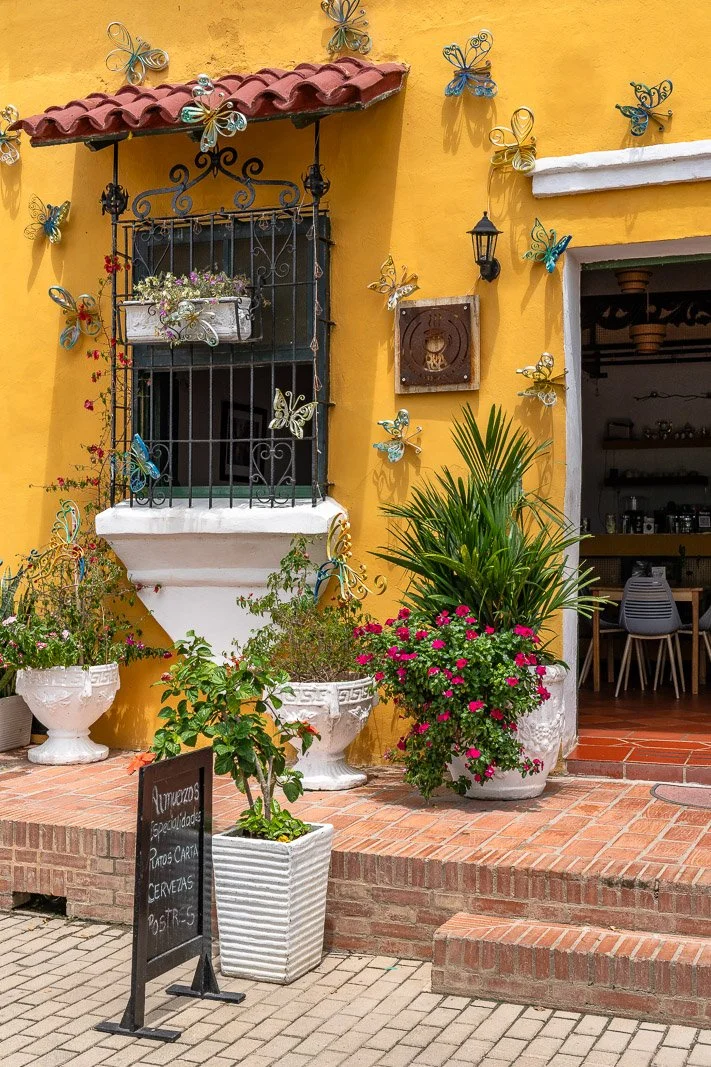  Gelbe Hausfassade in Santa Cruz de Mompox mit Metall-Schmetterlingen, Blumenarrangements und Blick in ein kleines Café. 