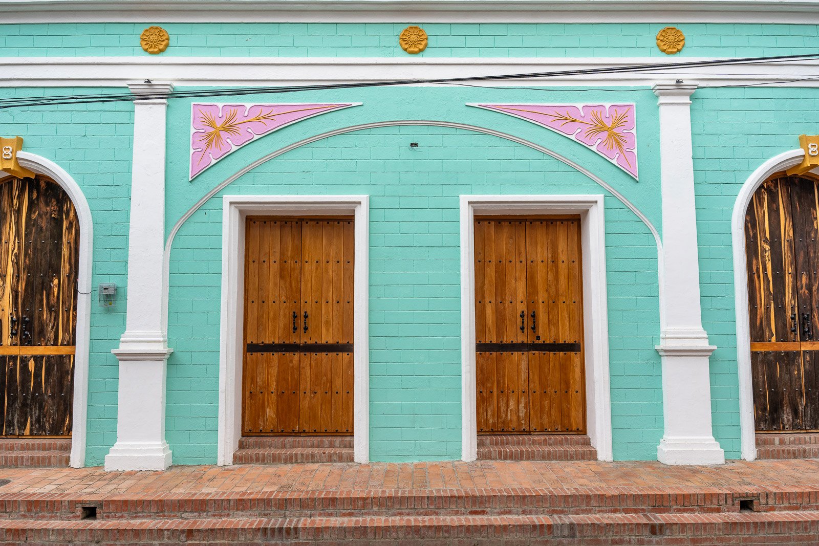  Türkisfarbene Kolonialfassade in Mompox mit zwei hölzernen Eingangstüren und pinken ornamentalen Elementen oberhalb der Bögen. 
