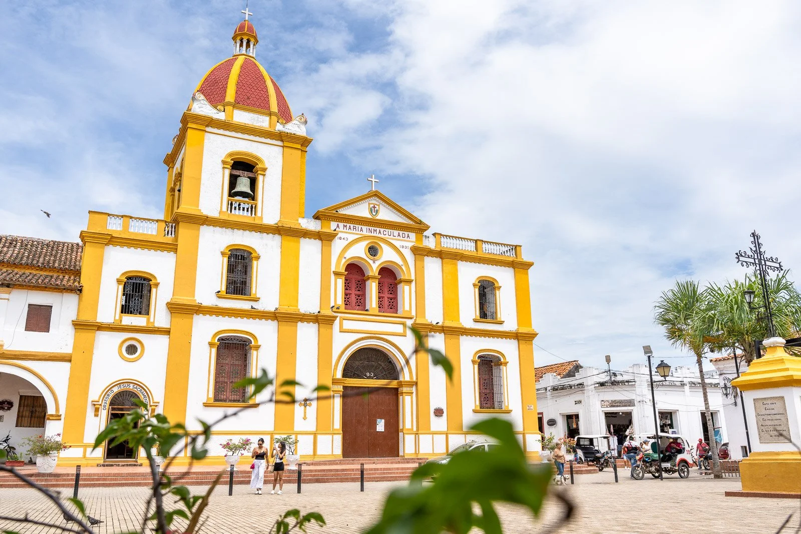  Fassade der Iglesia de la Concepción in Santa Cruz de Mompox, mit gelb-weißen Kolonialelementen und Menschen auf dem Platz. 