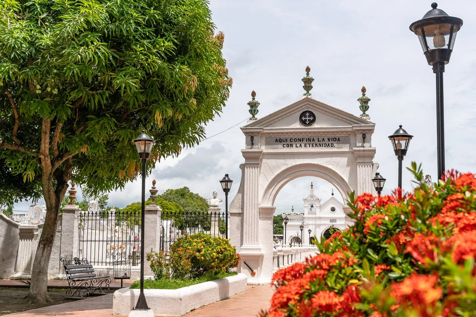  Monumentales Eingangsportal des Friedhofs von Mompox mit der Inschrift „Aquí confina la vida con la eternidad“, umgeben von Gartenanlagen und Laternen. 