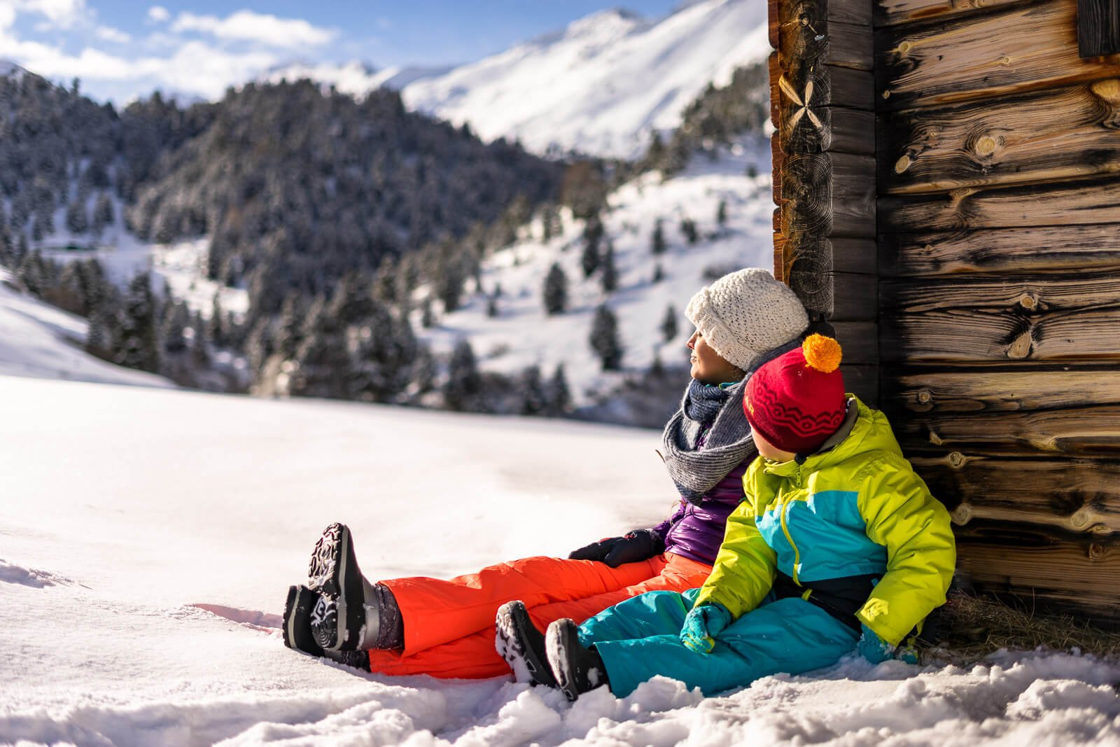 Mutter und Sohn sitzen in der Sonne an einer Holzhütte im Schnee und genießen den Winterurlaub in den Bergen.