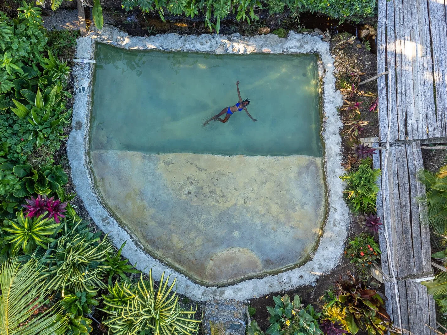 Drohnenaufnahme eines natürlichen Steinbeckens inmitten tropischer Pflanzen, Gast schwebt entspannt im Wasser.