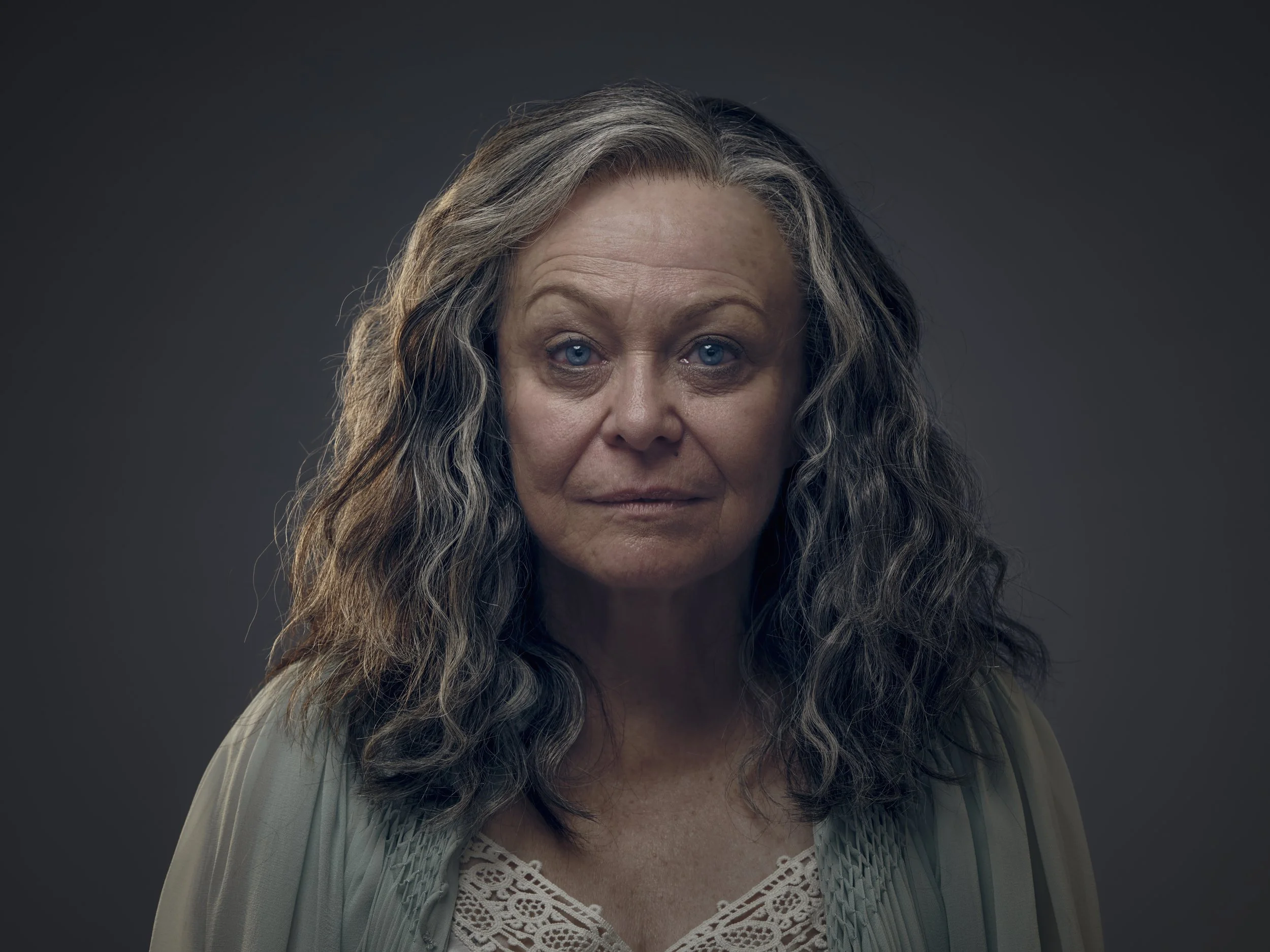 Close-up portrait of an older woman with long, wavy gray hair and blue eyes, wearing a light-colored blouse with lace detail, against a dark background.