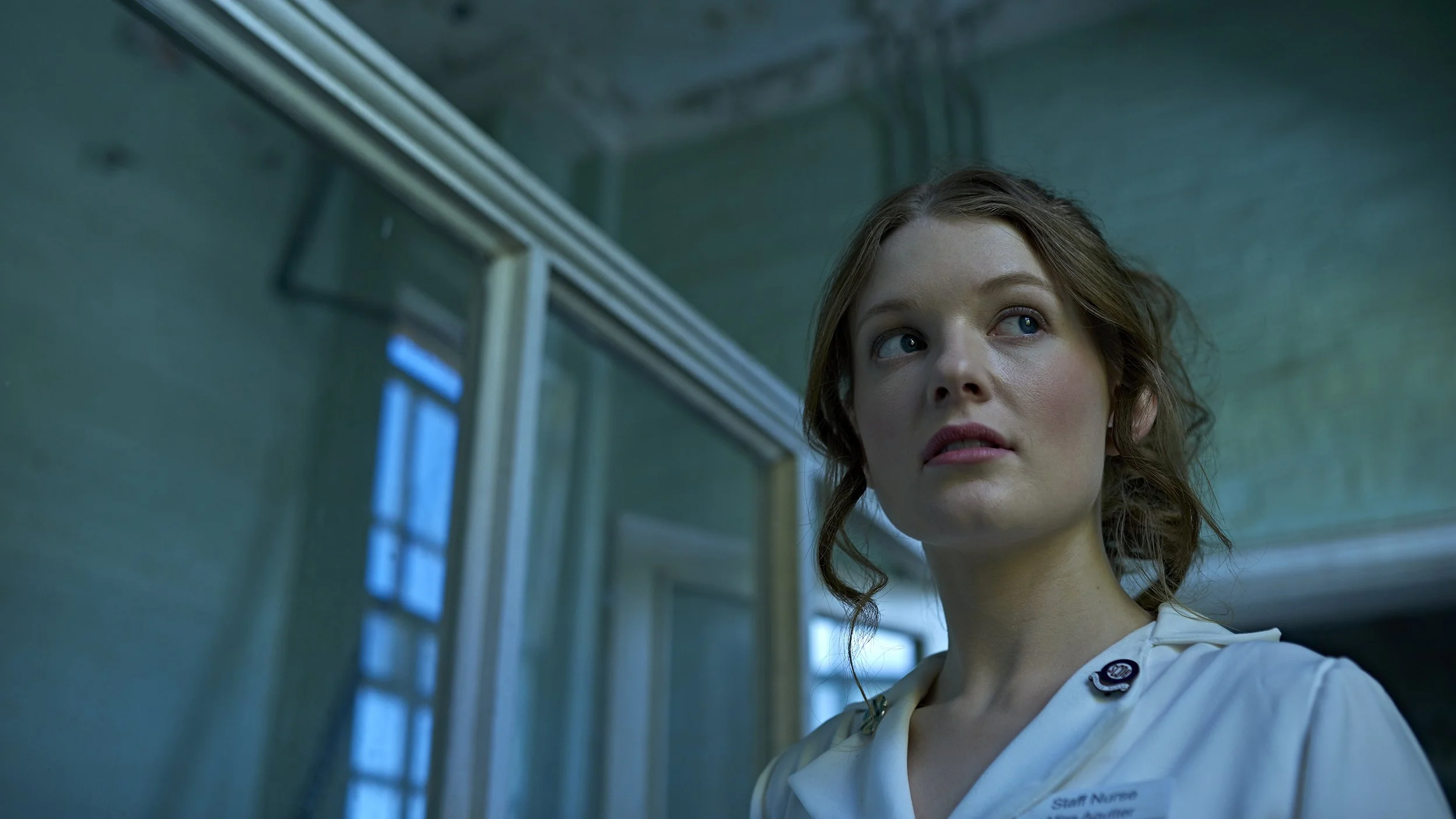 A young woman with curly brown hair, blue eyes, and light skin, looking to the side, standing indoors near a wall with windows, wearing a white uniform with a name tag.