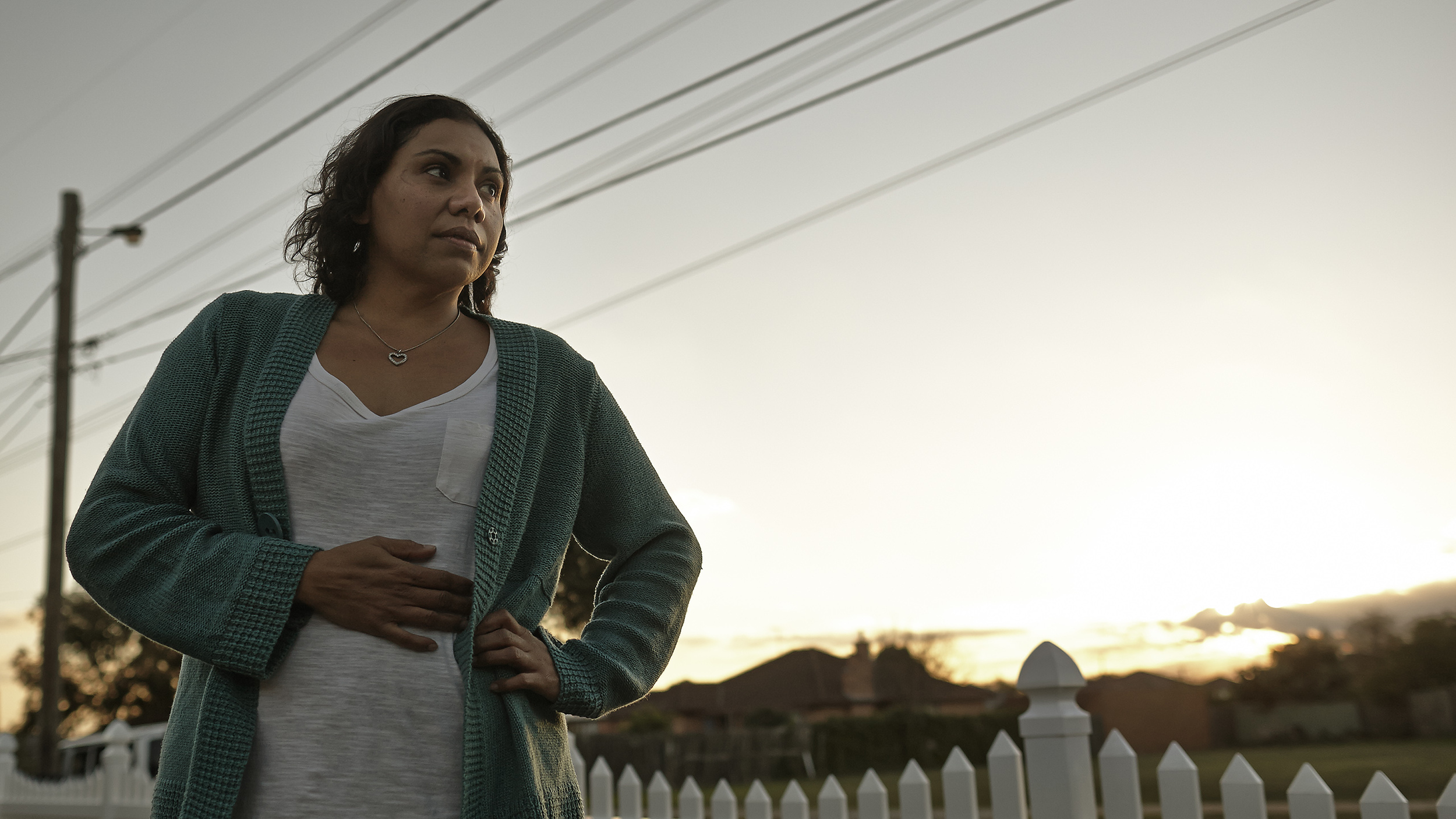 A woman wearing a green cardigan and white top, standing outdoors during sunset, with her hand on her stomach, looking thoughtful.