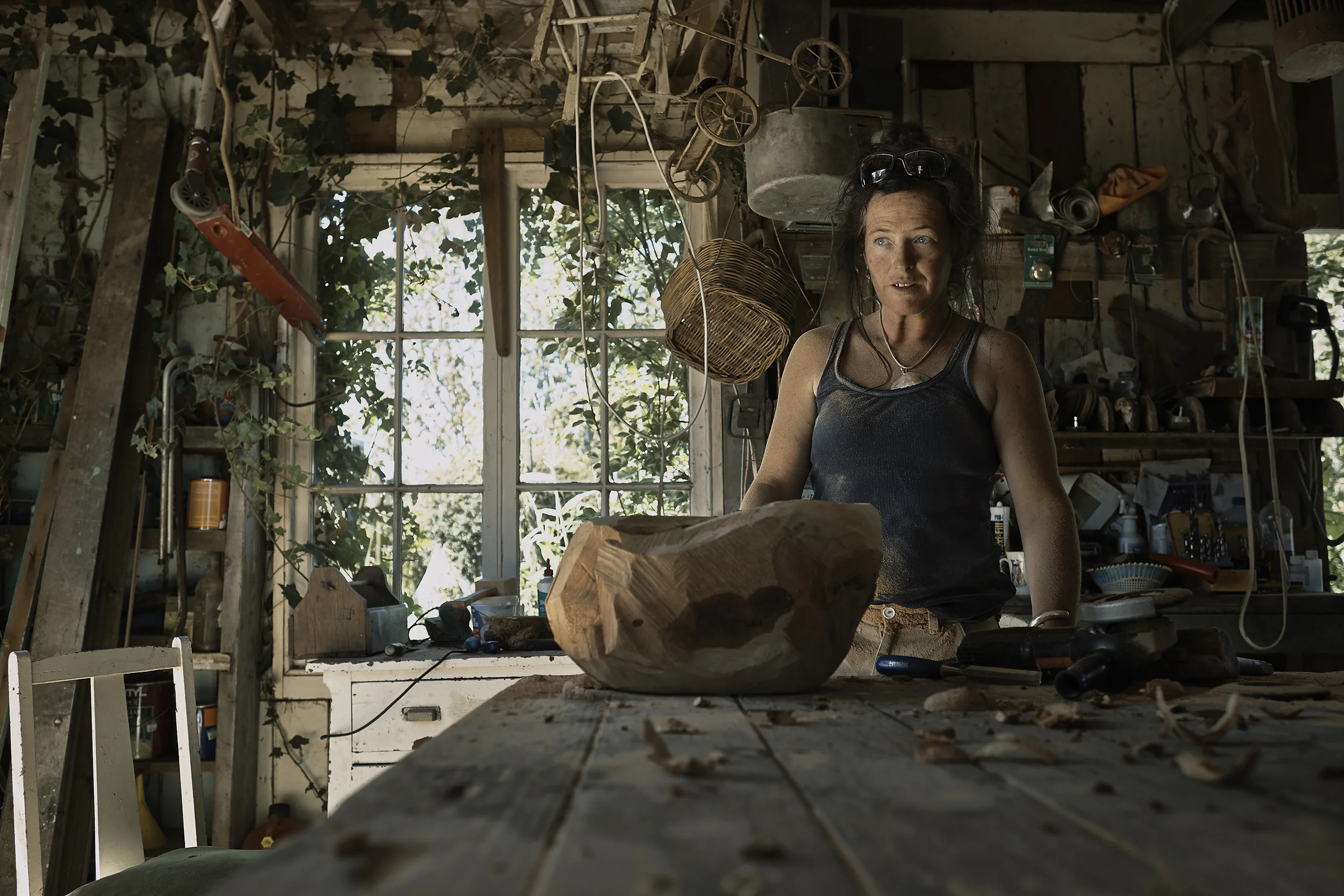Woman with black curly hair wearing a black tank top and sunglasses on her head, standing in a cluttered woodworking workshop with wood shavings and tools, lit by natural light through a large window background.