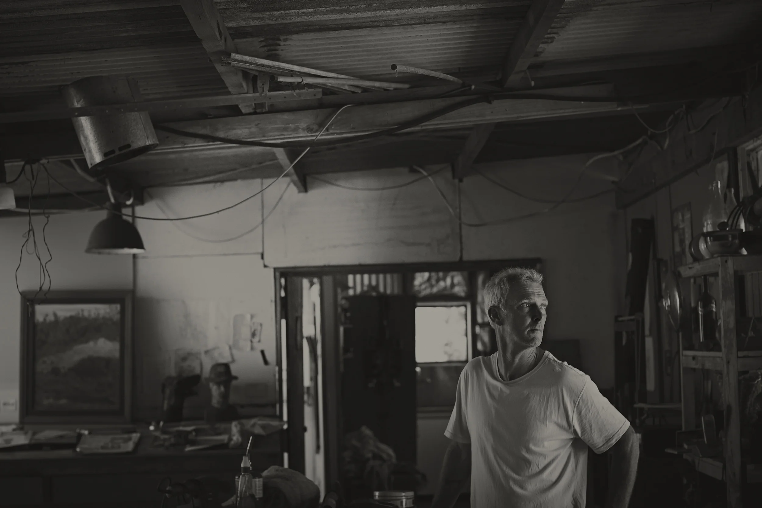 A man with gray hair wearing a white t-shirt in a workshop with a cluttered wooden ceiling and various tools and objects around him.