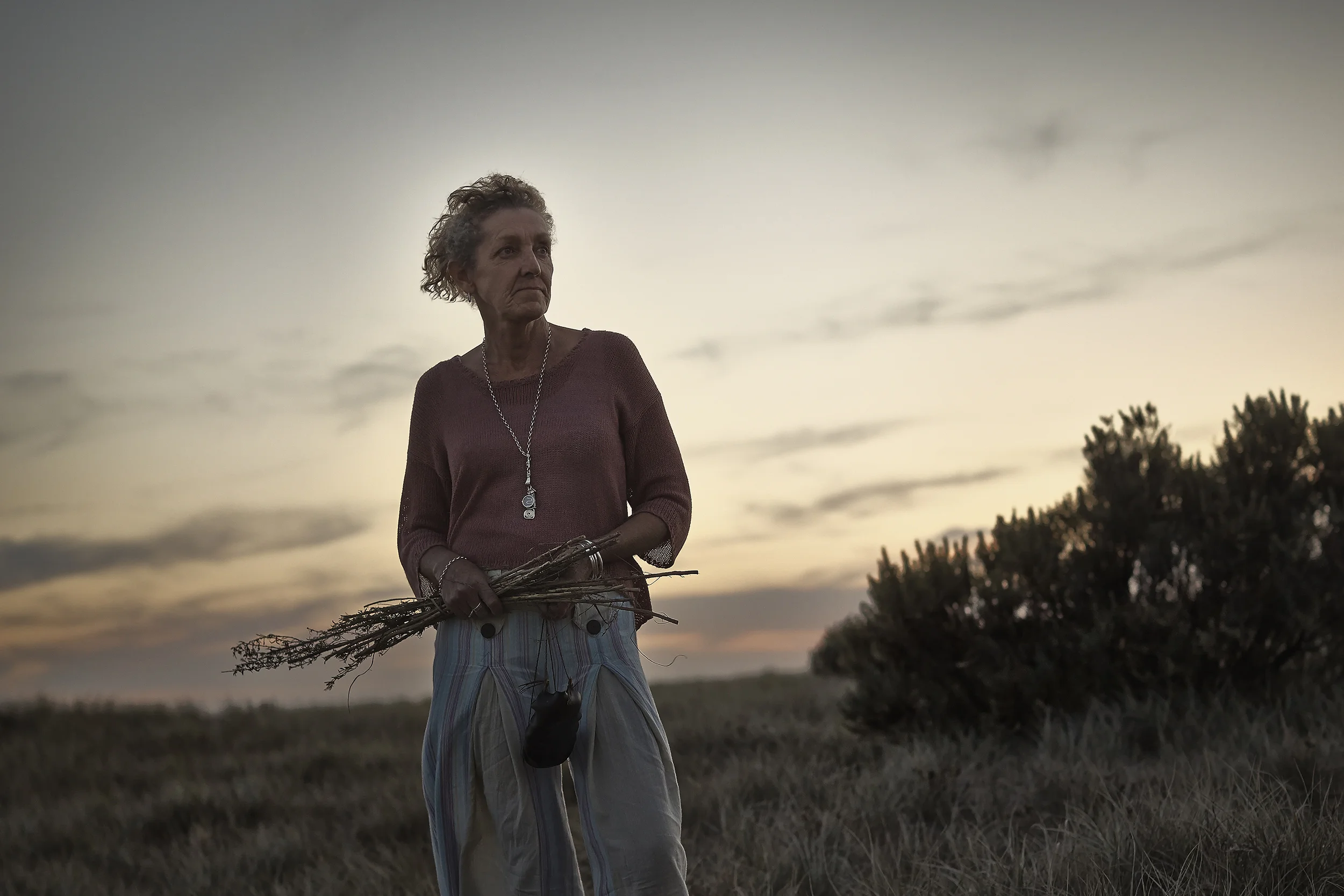 An elderly woman with curly gray hair standing outdoors during sunset, holding a bouquet of sticks, wearing a long-sleeved burgundy sweater and patterned pants, with a necklace, in a field with trees in the background.