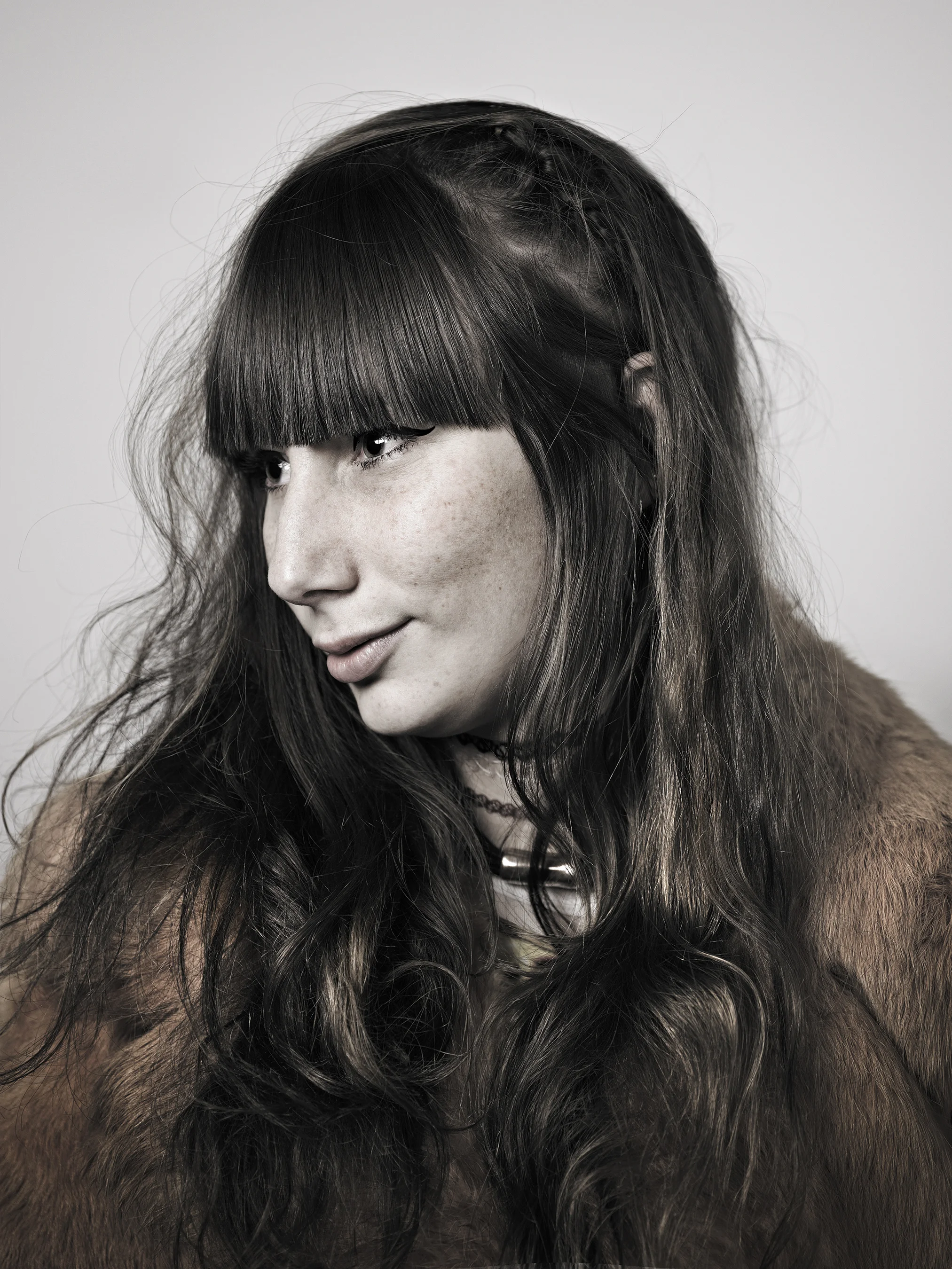 A woman with straight black hair and bangs, wearing a brown fur coat, looking down and to the side against a plain gray background.
